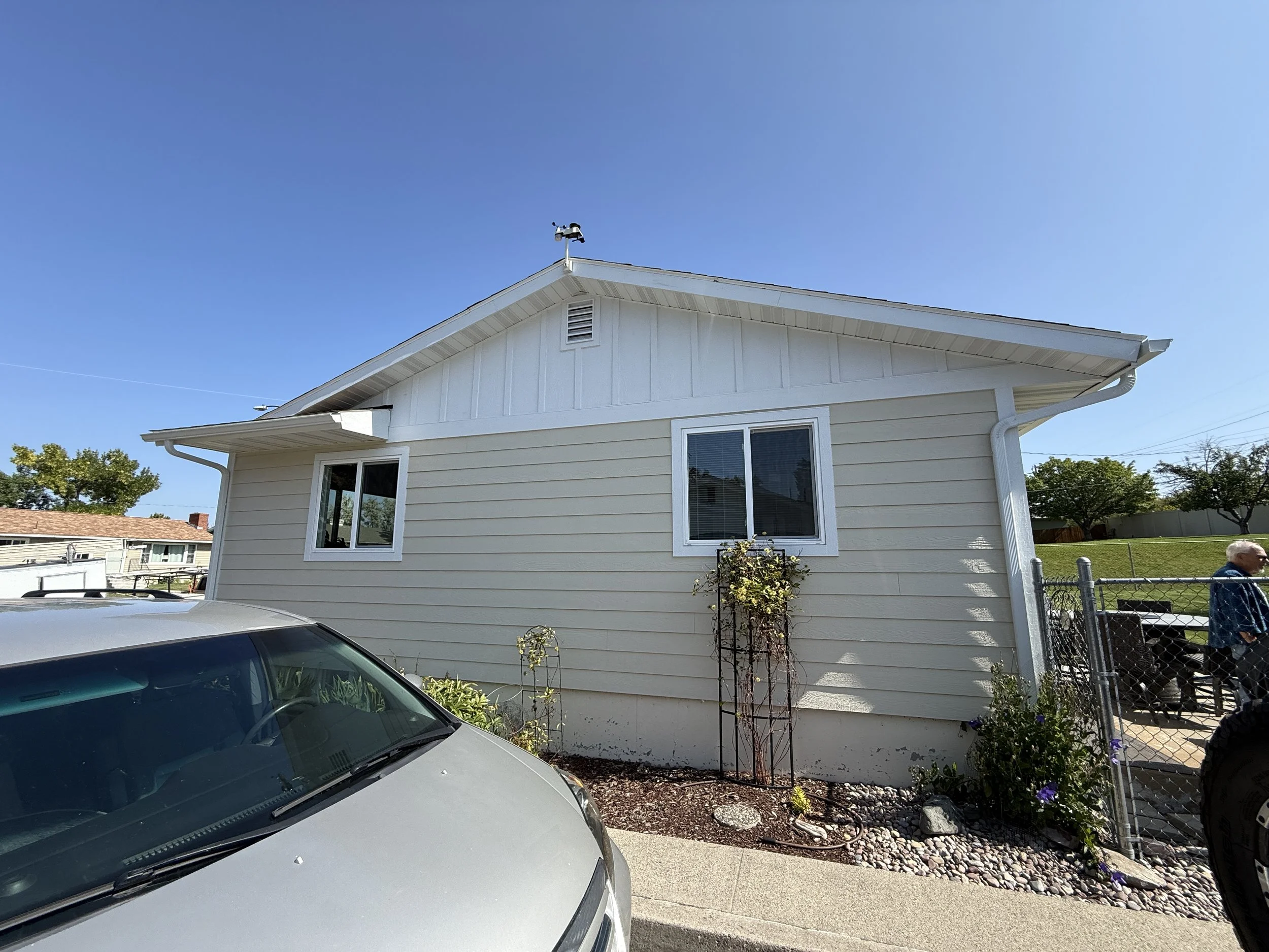 Front view of a beige house with white trim, two windows, a small garden with plants, and a car parked in the driveway on a clear sunny day.