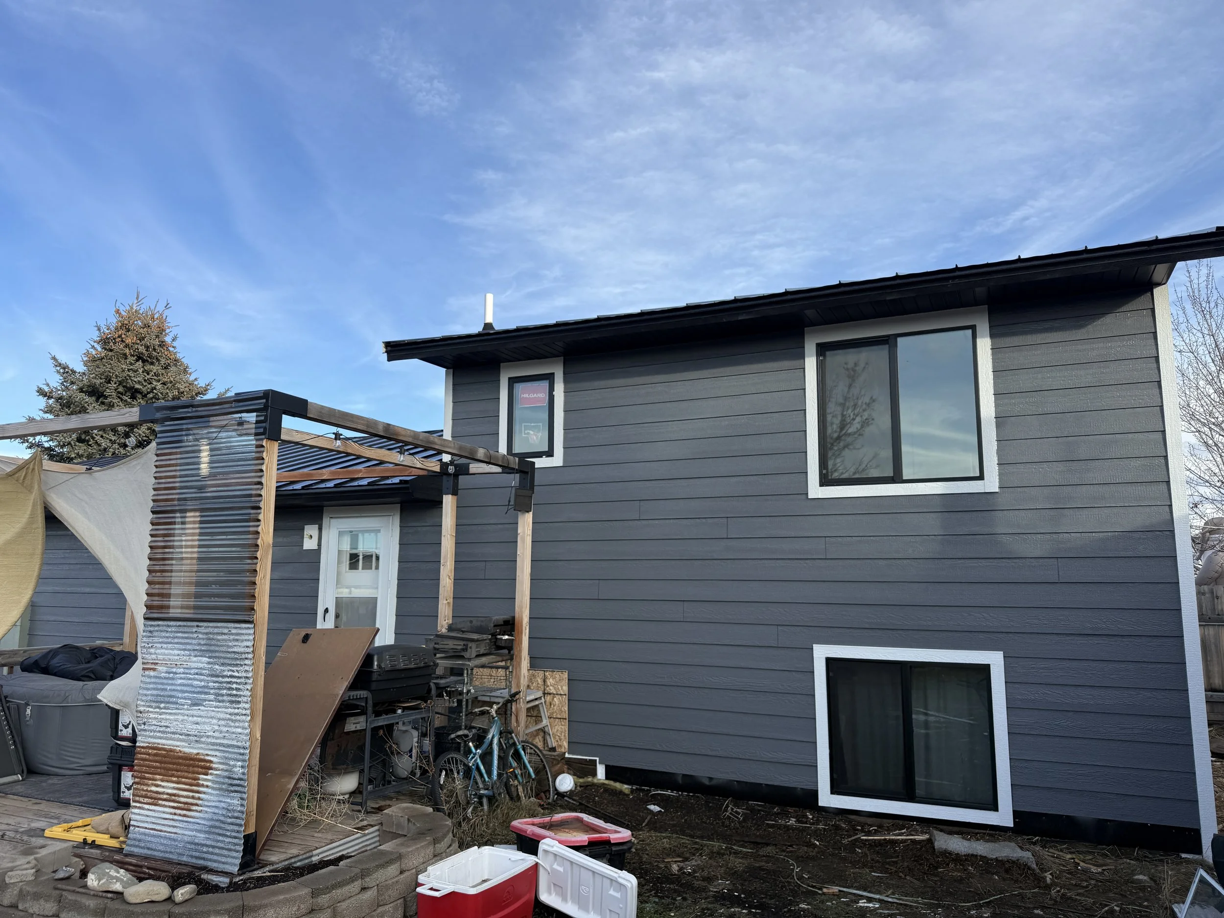 A two-story gray house with white window frames, a sliding glass door, a small window, and a backyard with a construction or storage area containing equipment, a bicycle, and miscellaneous materials.