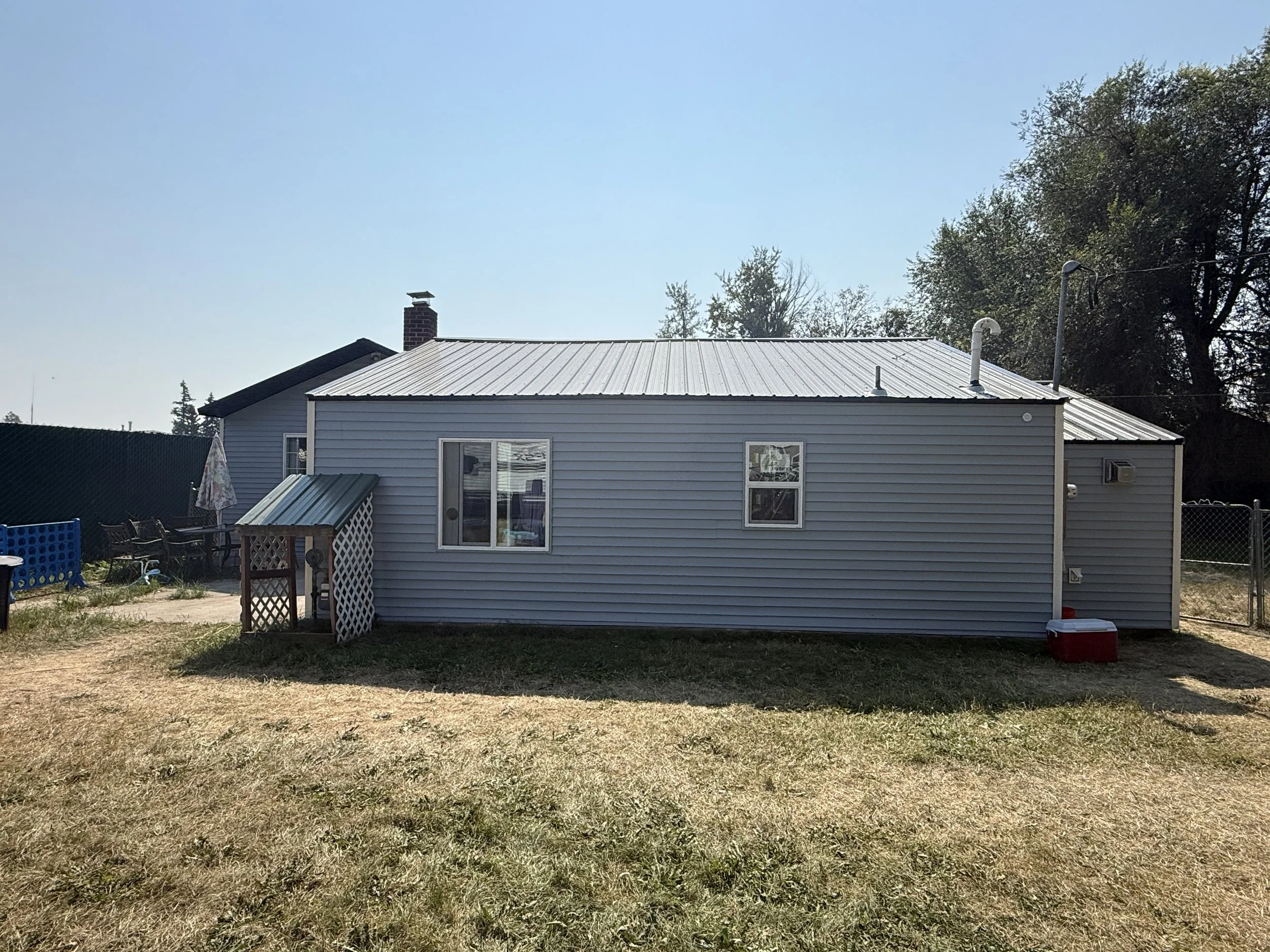 Backyard view of a house with gray siding, metal roof, and small windows, with outdoor furniture and a small shed on the side.
