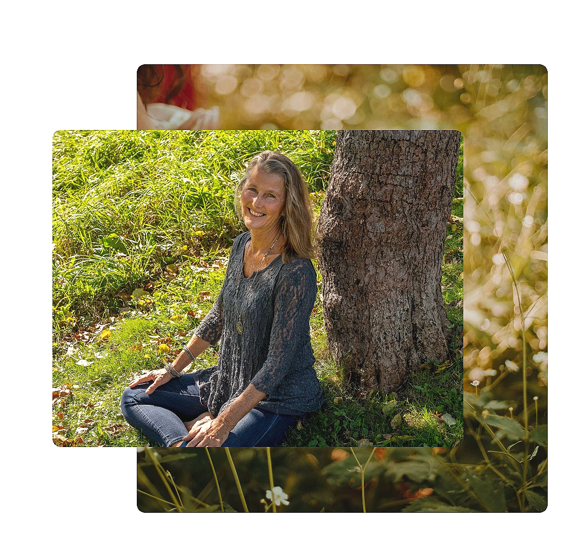 A smiling woman with blonde hair sitting on grass next to a large tree in a sunny outdoor setting with green foliage.
