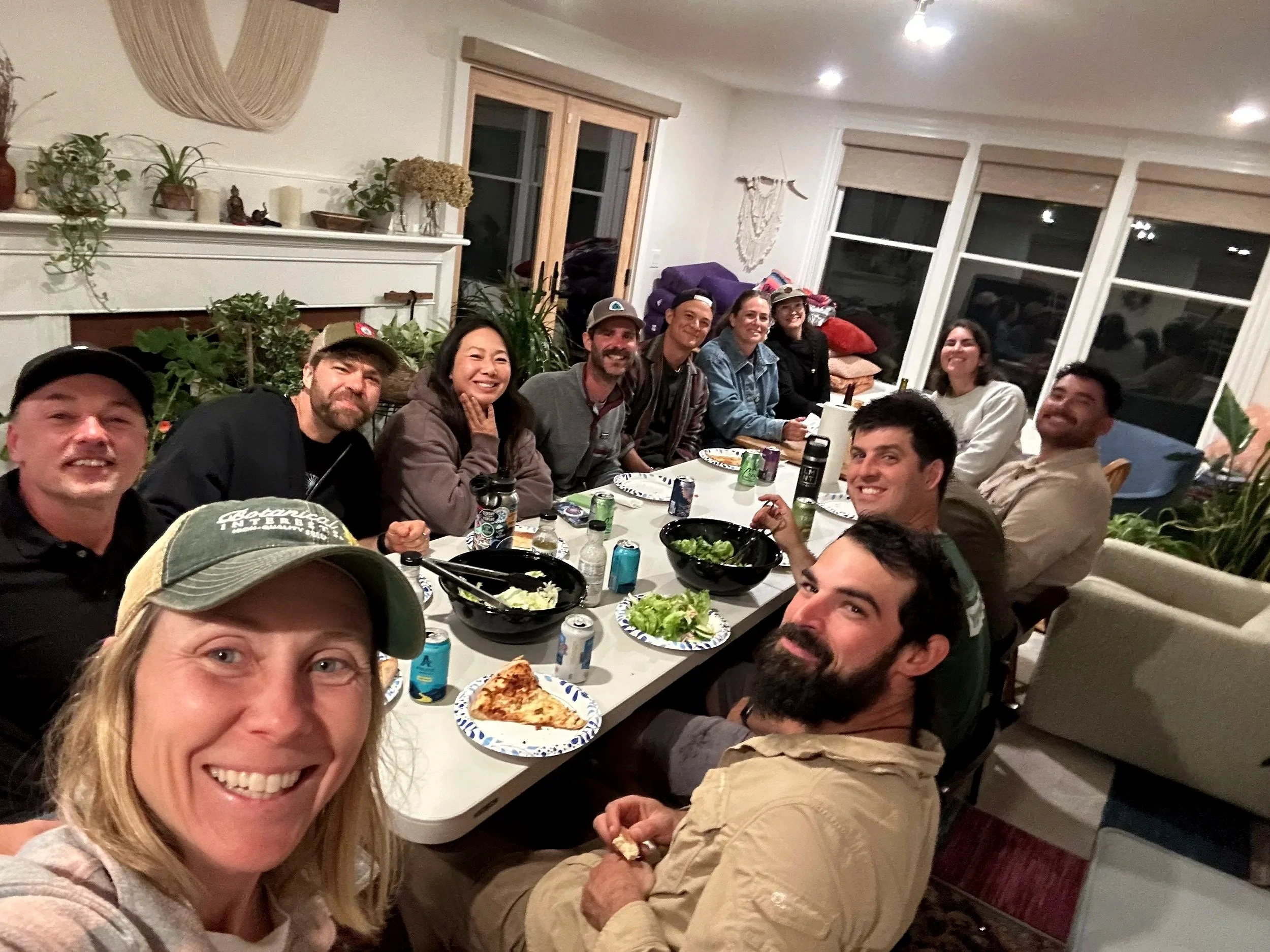A group of people sitting around a dining table at a house party, smiling for the camera, with food and drinks on the table, and home decor visible in the background.