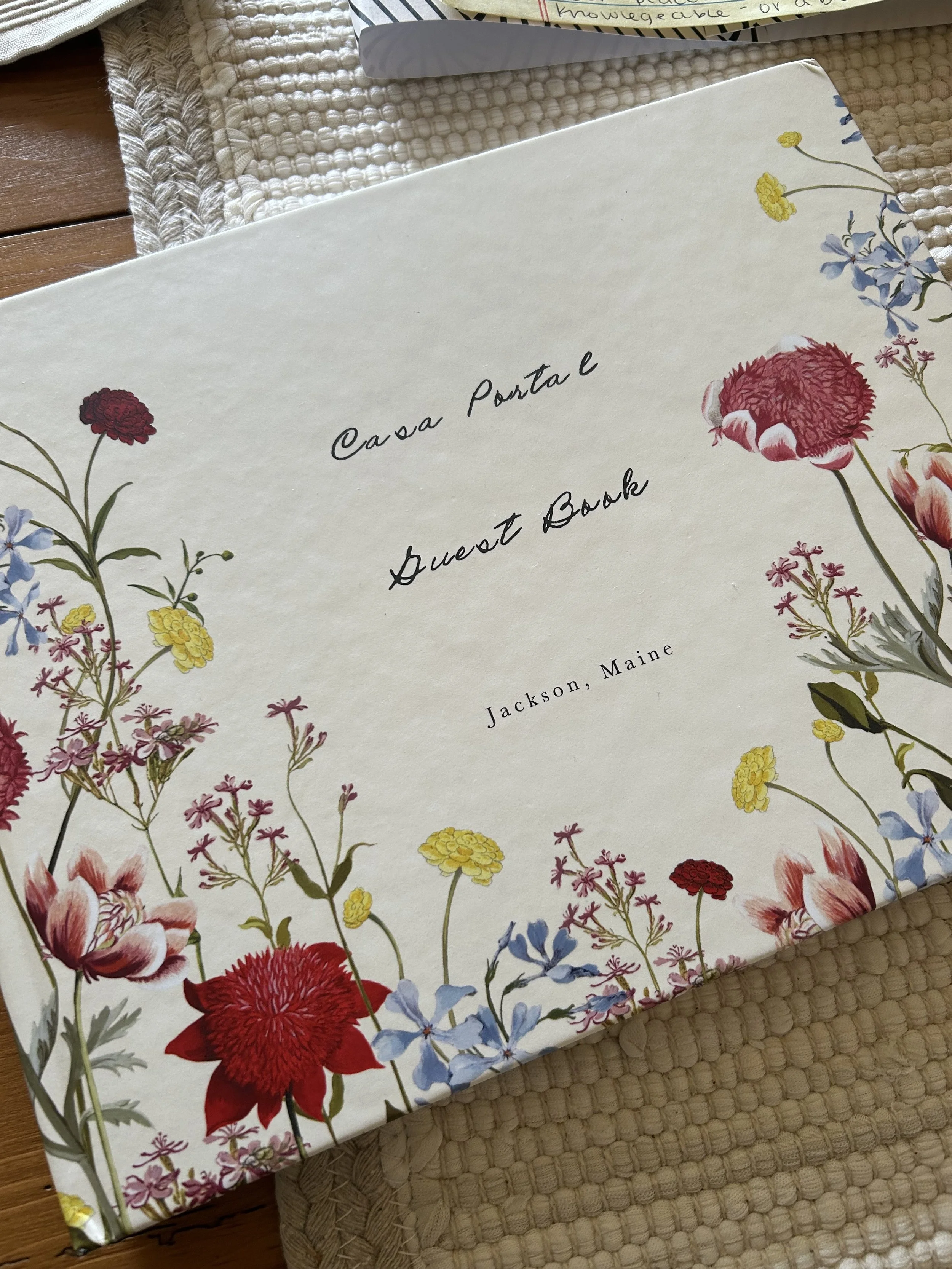 A decorative book titled 'Casa Portal guest book' lying on a beige woven placemat on a wooden surface, surrounded by colorful flowers on the cover.