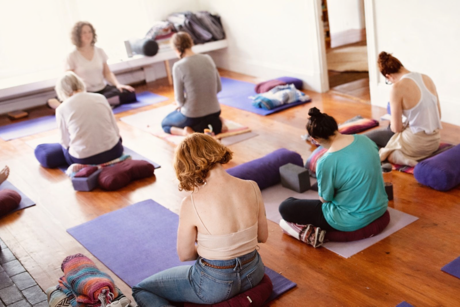 Group of women participating in a yoga or meditation class indoors, sitting on cushions and mats in a circle, with sunlight coming through a window.