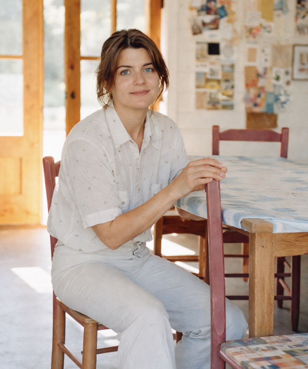 A woman sitting on a wooden chair at a dining table, wearing a white shirt and cream-colored pants, inside a room with wooden walls and large windows.