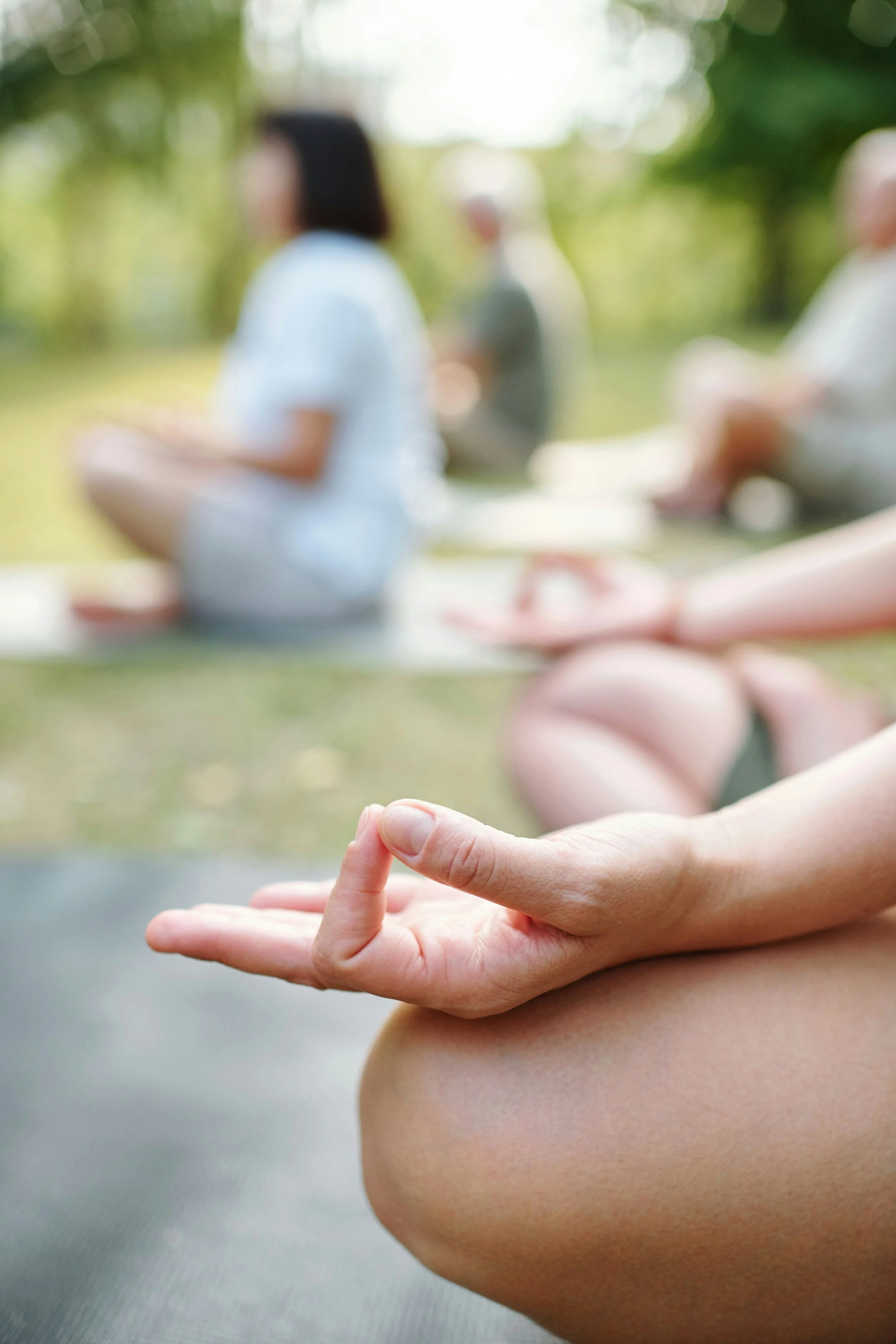 Close-up of a person practicing yoga in a seated meditation pose in an outdoor setting with blurry people sitting on yoga mats in the background.