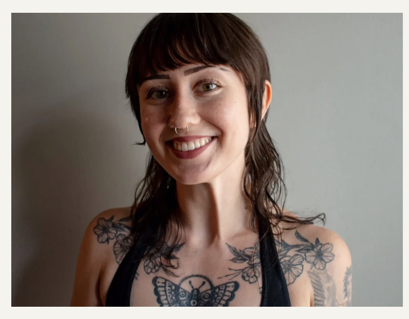 Smiling woman with short brown hair and tattoos on her shoulders and chest, wearing a black tank top.
