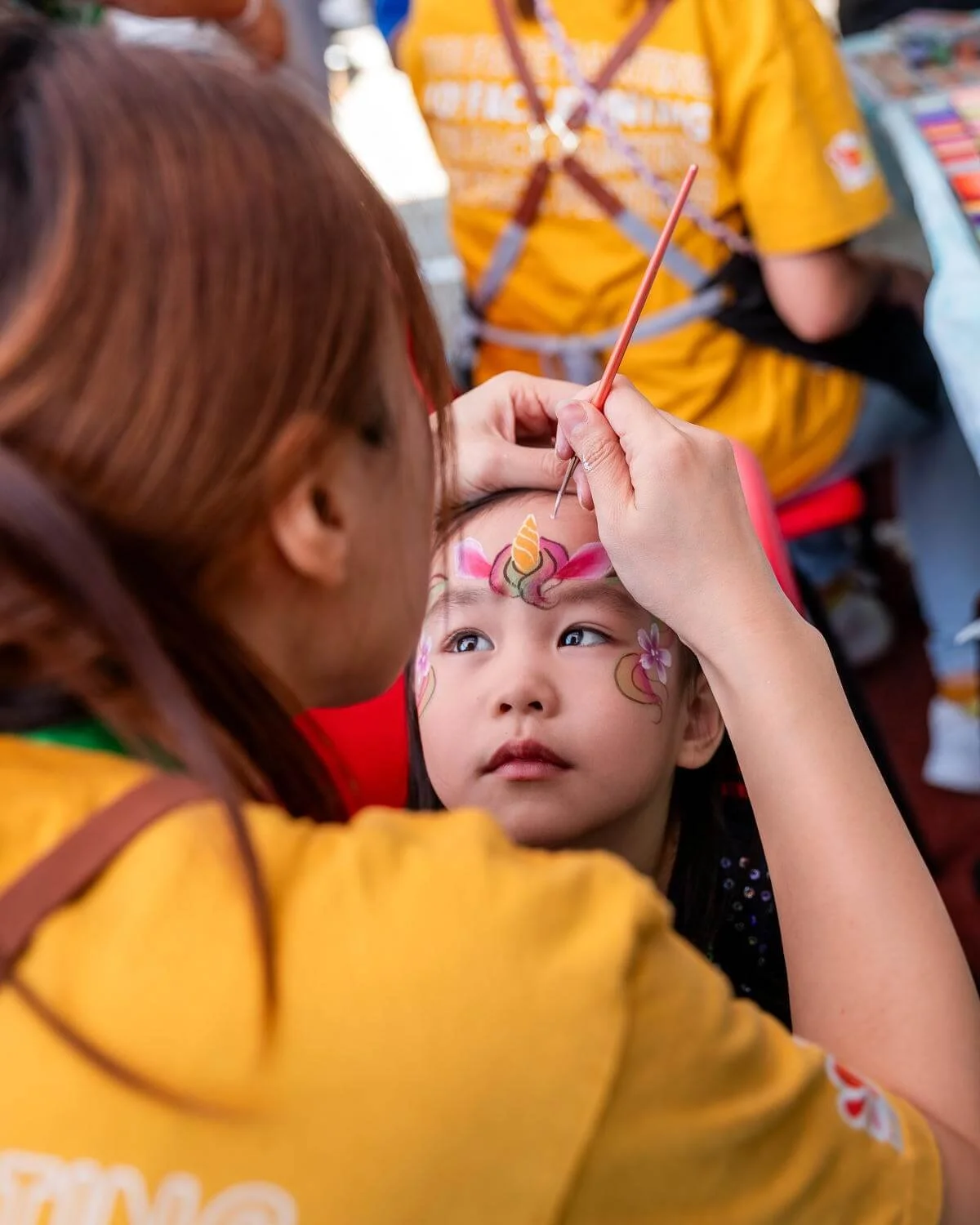 A girl with face paint designed like a unicorn with flowers is getting her face painted by an artist at a festival or fair.