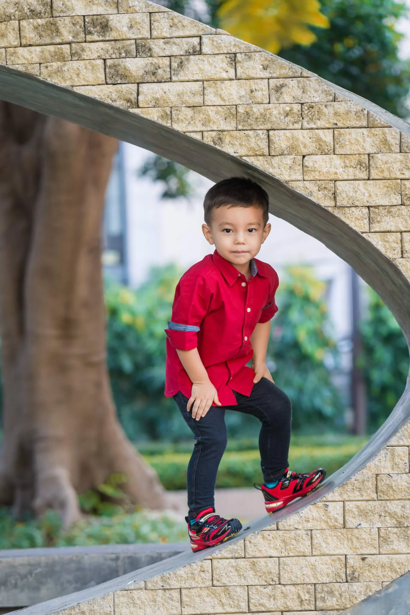 A young boy in a red shirt and black pants climbing a stone structure in a park, with greenery in the background.