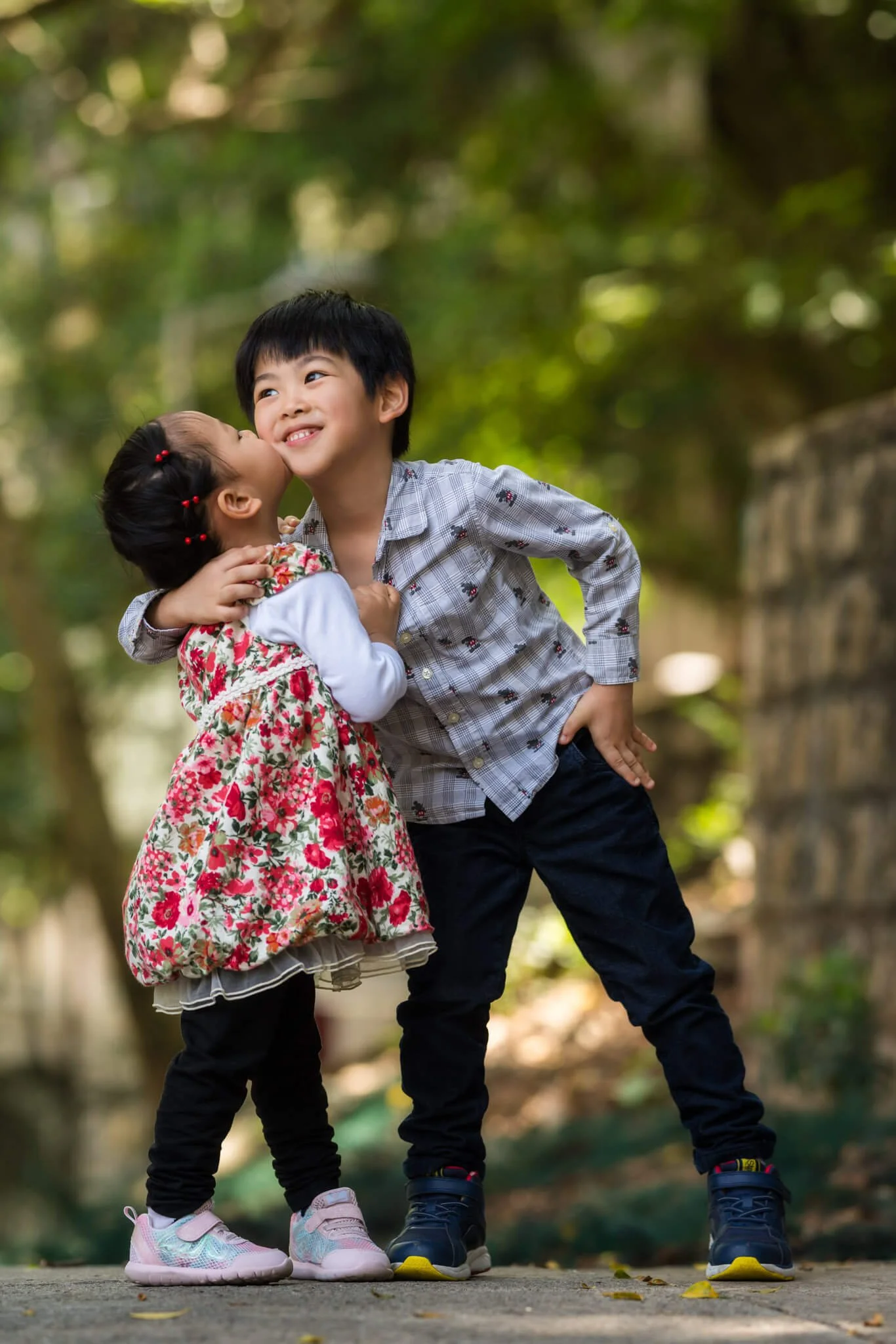 Two young children, a girl and a boy, outdoors in a park. The girl is wearing a floral dress and kissing the boy on the cheek. The boy is smiling and wearing a plaid shirt.