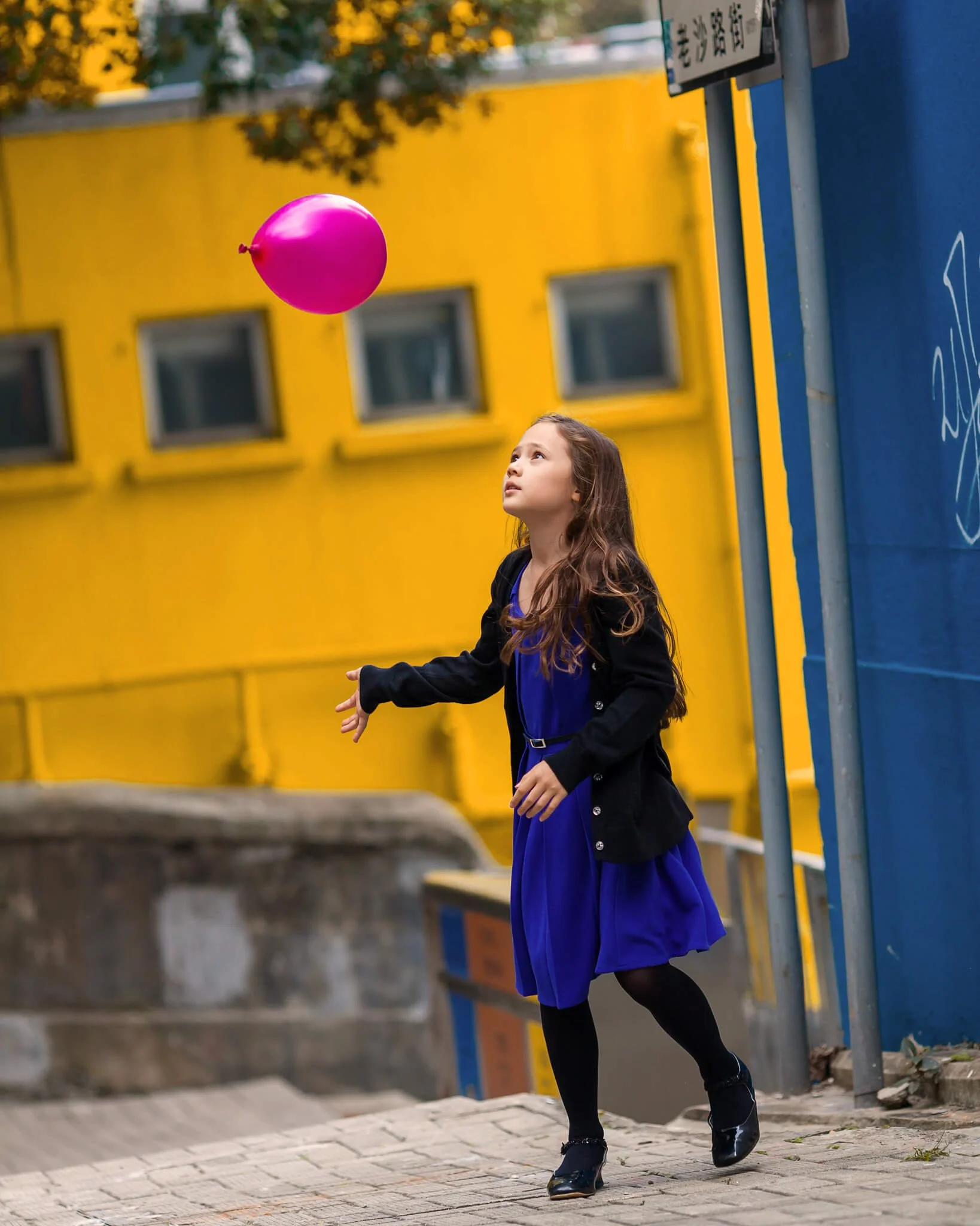 Girl in purple dress playing with pink balloon in urban area with colorful buildings.