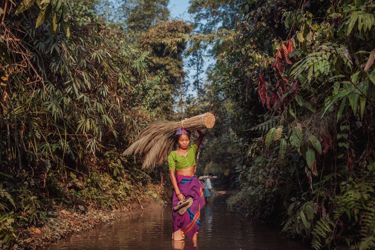 Chakma woman stands in a stream with brooms for sale on her head. 
