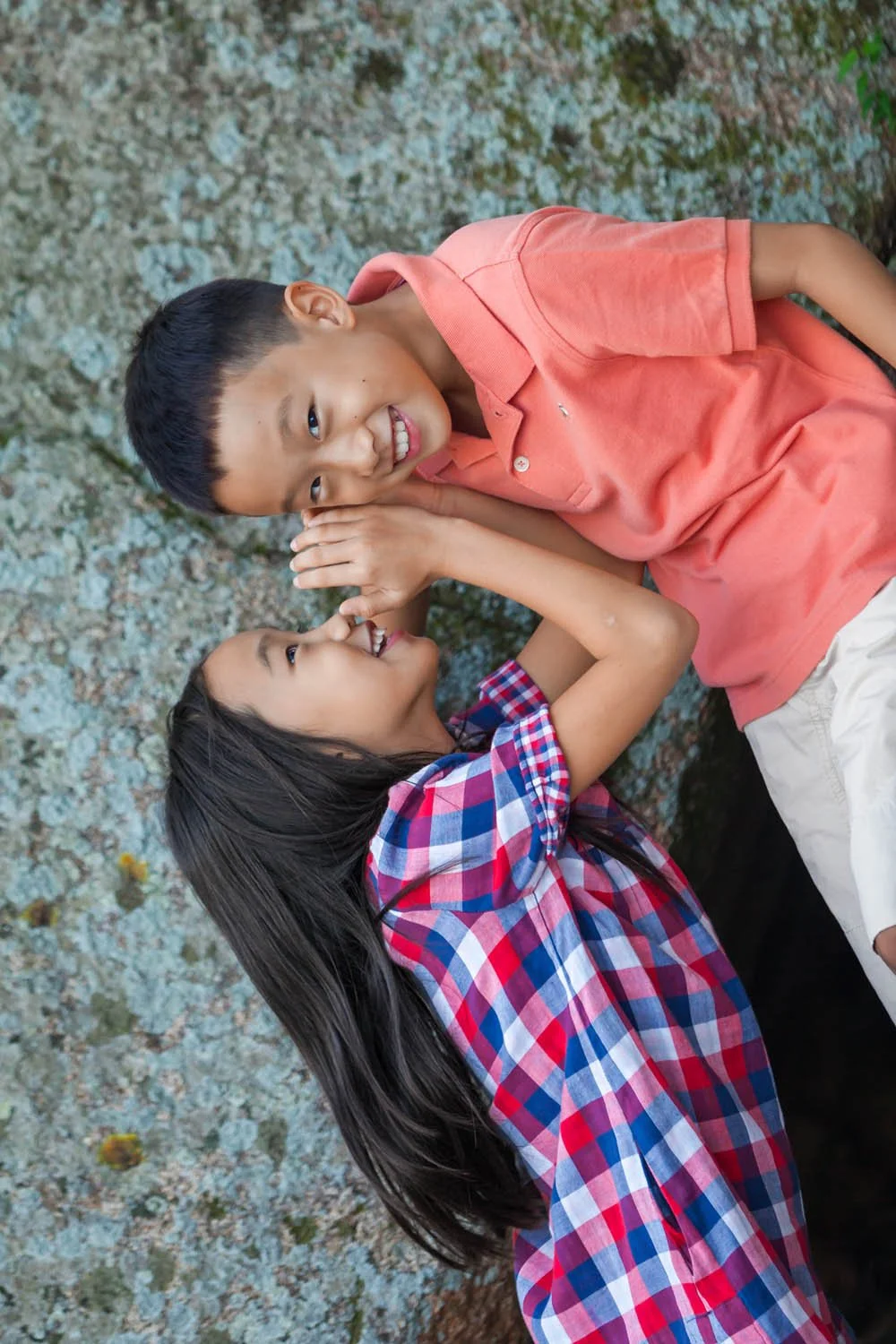 Two children lying on a stone surface; one is whispering to the other. The boy in a coral shirt is smiling, and the girl in a plaid shirt is whispering with a playful expression.