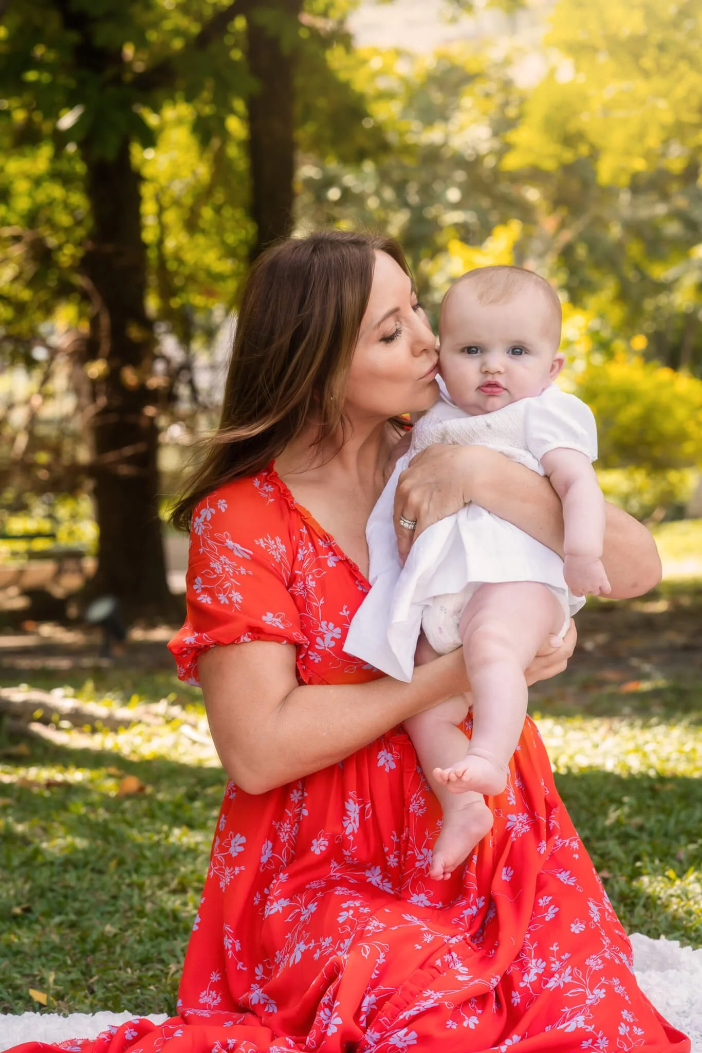 A woman in a red floral dress sitting on grass in a park, holding and kissing a baby wearing a white outfit.