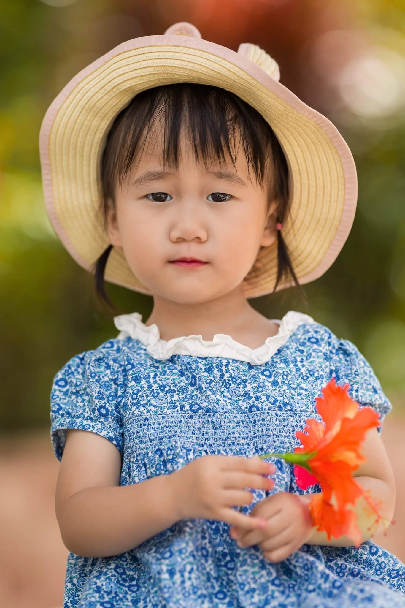 Young child wearing a blue floral dress and straw hat, holding a bright orange flower during a photo shoot