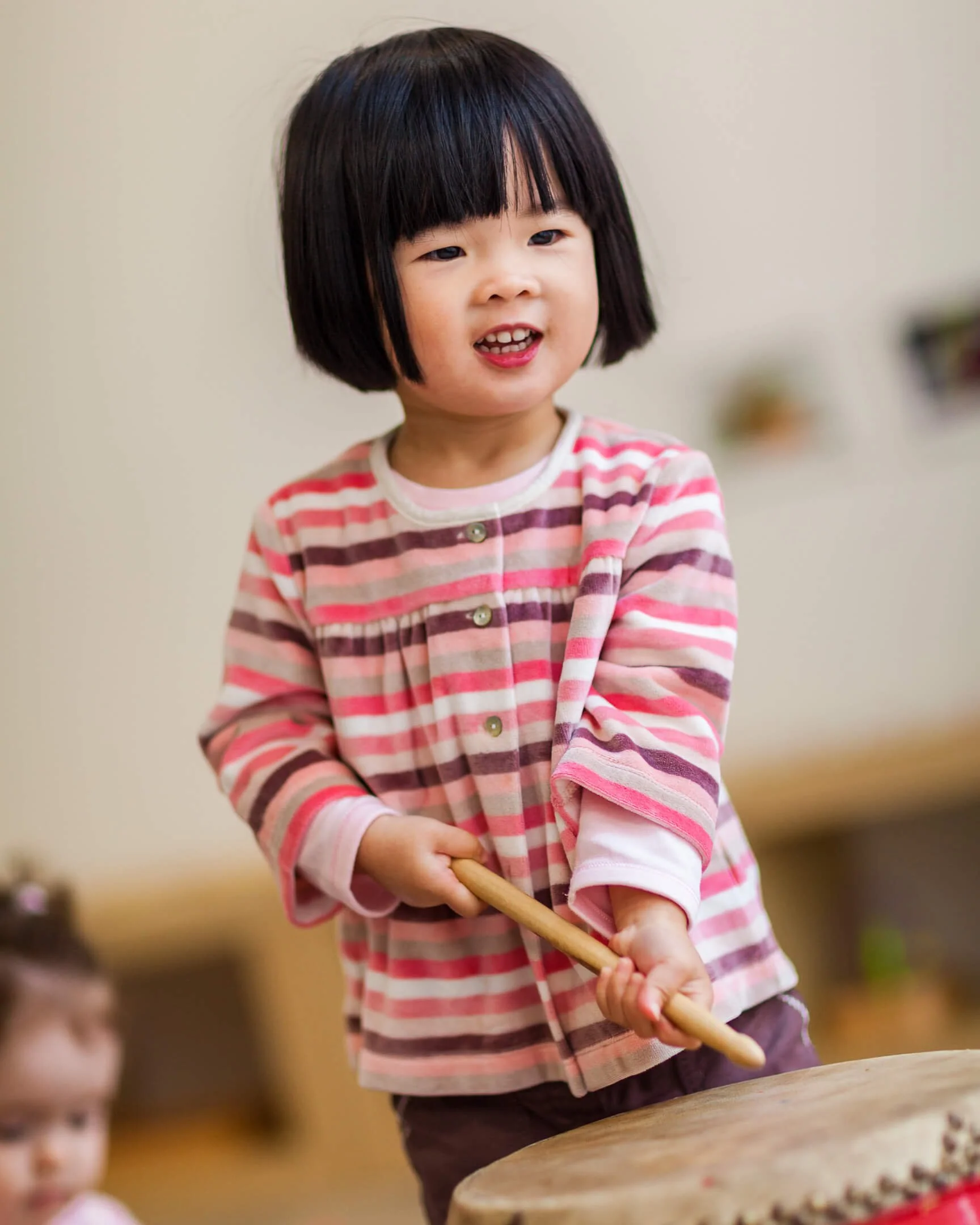 Young child in a striped shirt playing a drum with a stick.
