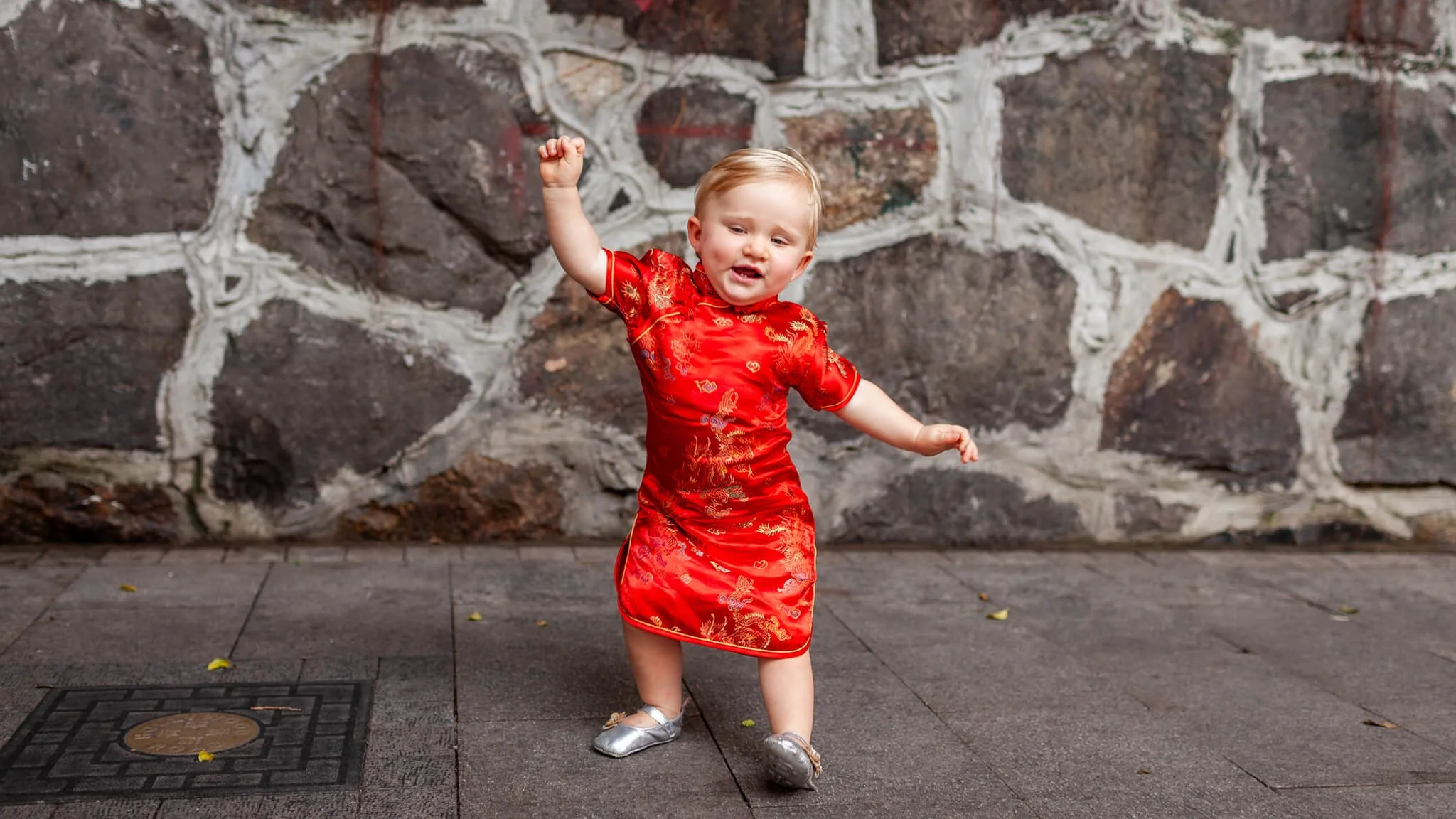A young toddler in a red traditional Chinese dress dancing in front of a stone wall