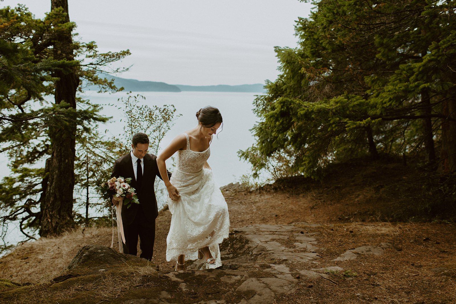 Bride and groom hike up a small cliffside with the Puget Sound in the backdrop