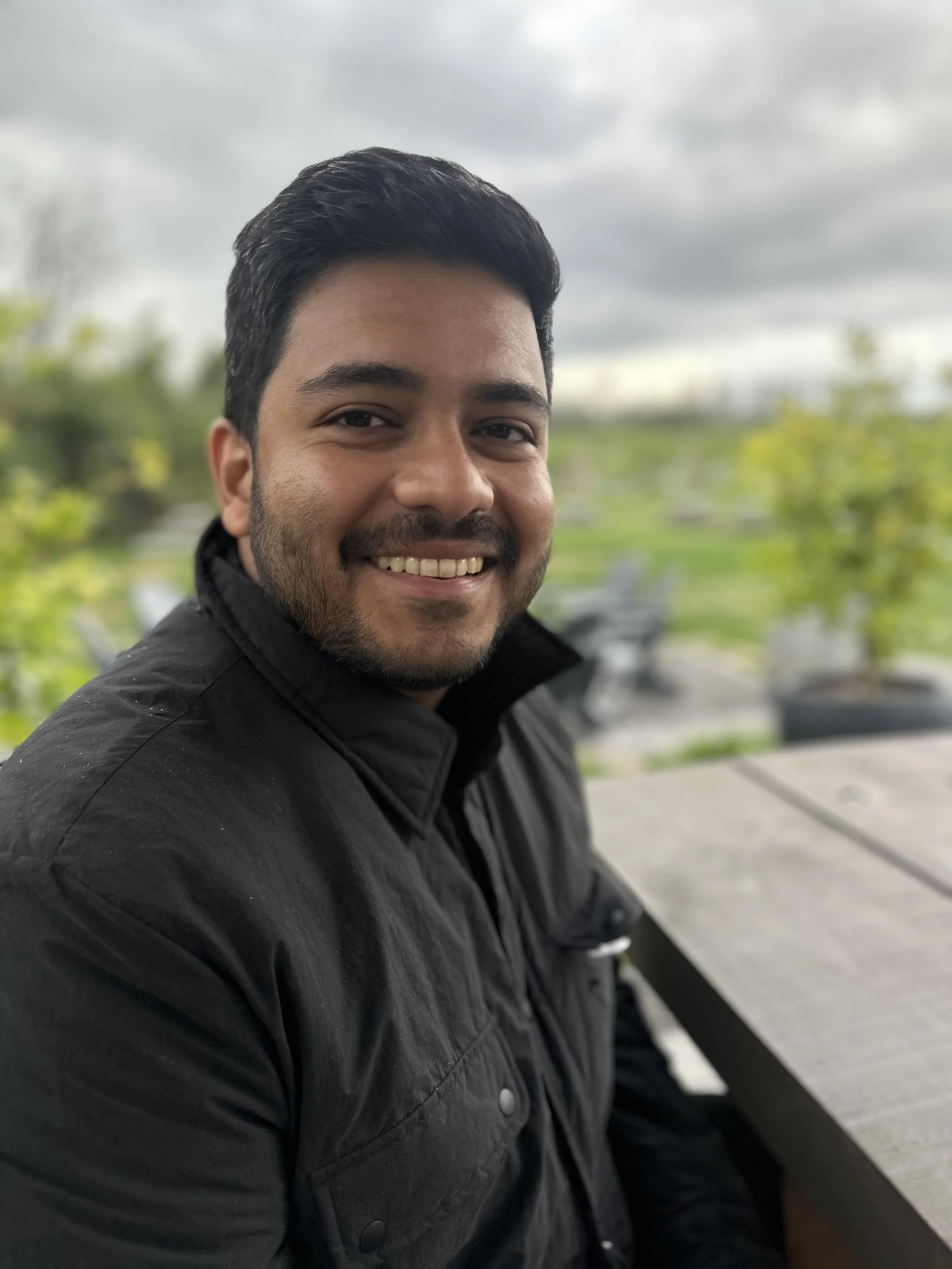 Please read AI's description of this picture of me in PEC: a man with short black hair, a beard, and a big smile, wearing a black jacket, sitting outside at a wooden picnic table on a cloudy day with greenery and overcast sky in the background.