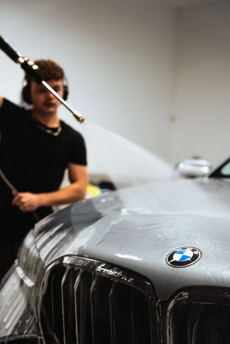 Technician in ear defenders pressure rinsing the hood of a silver BMW X6 during a detail at Ascension Automotive