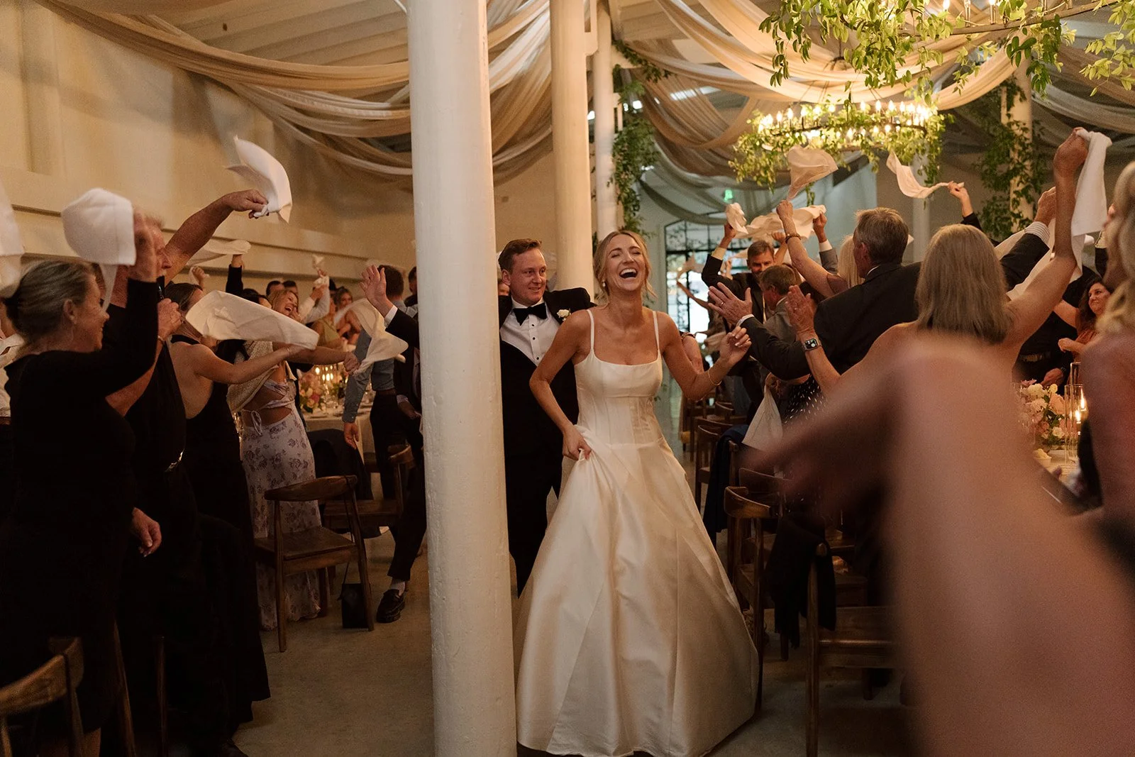 This is a photo of a smiling bride and groom. They are making their official entrance to their Reception! Their guests are standing at the tables cheering them on and waving napkins above their heads as the couple walks past!