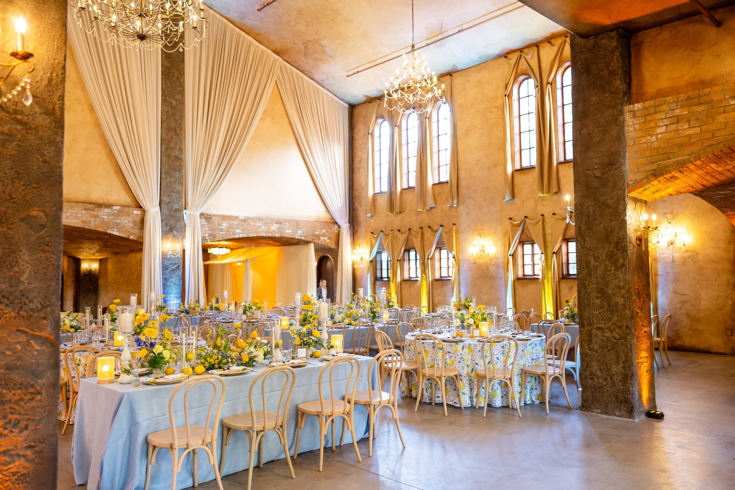This is a wedding reception space with high ceilings, several large windows lined with draping, and a chandelier hangs above. The room is full of natural light and uplighting has added pastel colored lights to the walls, matching the table decor.