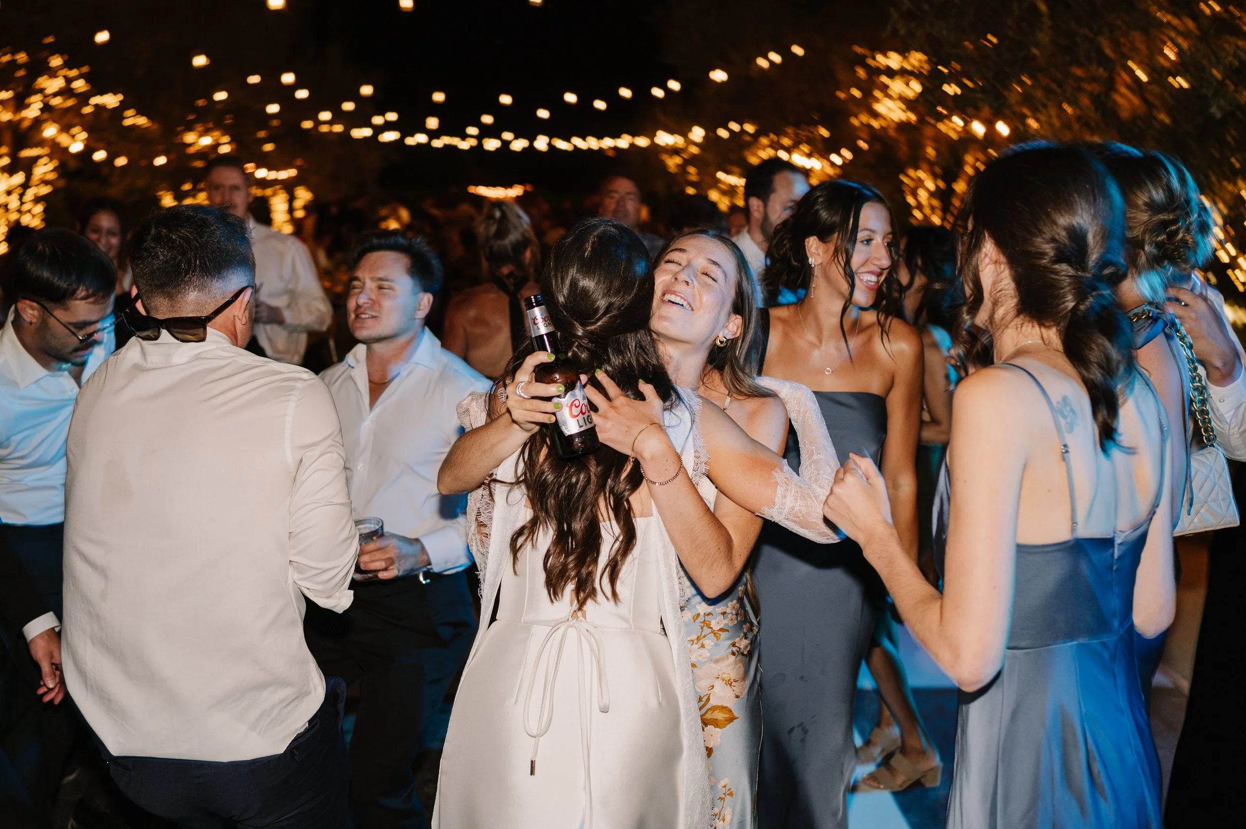 This is a photo of a bride surrounded by her loved ones on the dance floor. Everyone is smiling, dancing, and one of the bride's friends has pulled her in for a hug! The dance floor is outside, surrounded by trees, and bistro lights hang overhead.