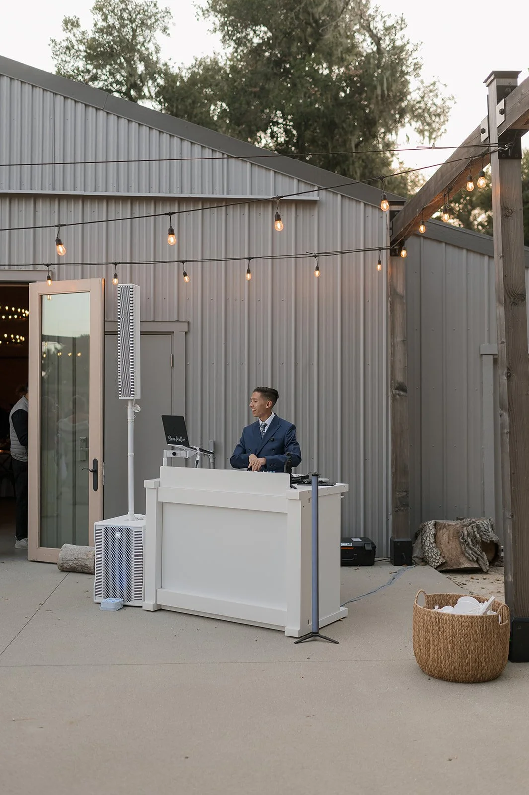DJ Steven stands behind his booth at an outdoor wedding in Templeton, CA. He's in front of a barn with gray siding, bistro lights hand above, and he's smiling in a blue suit.