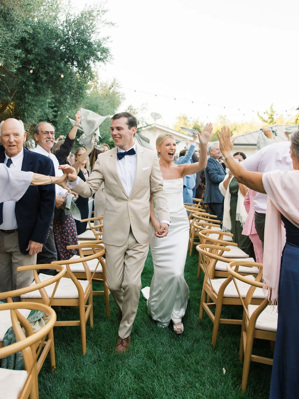 A bride and groom walk through an aisle of standing, cheering guests as they make their Grand Entrance to their Reception. The bride is giving a high five, the groom is shaking a hand, and everyone is smiling.