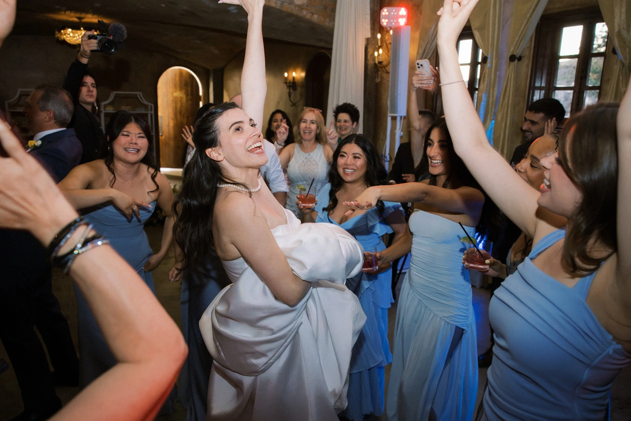 A bride is at the center of the dance floor, smiling with a hand in the air, as her friends cheer and dance with her. Most of her friends are wearing blue dresses and one guest is holding a phone, capturing a quick video of this fun moment!