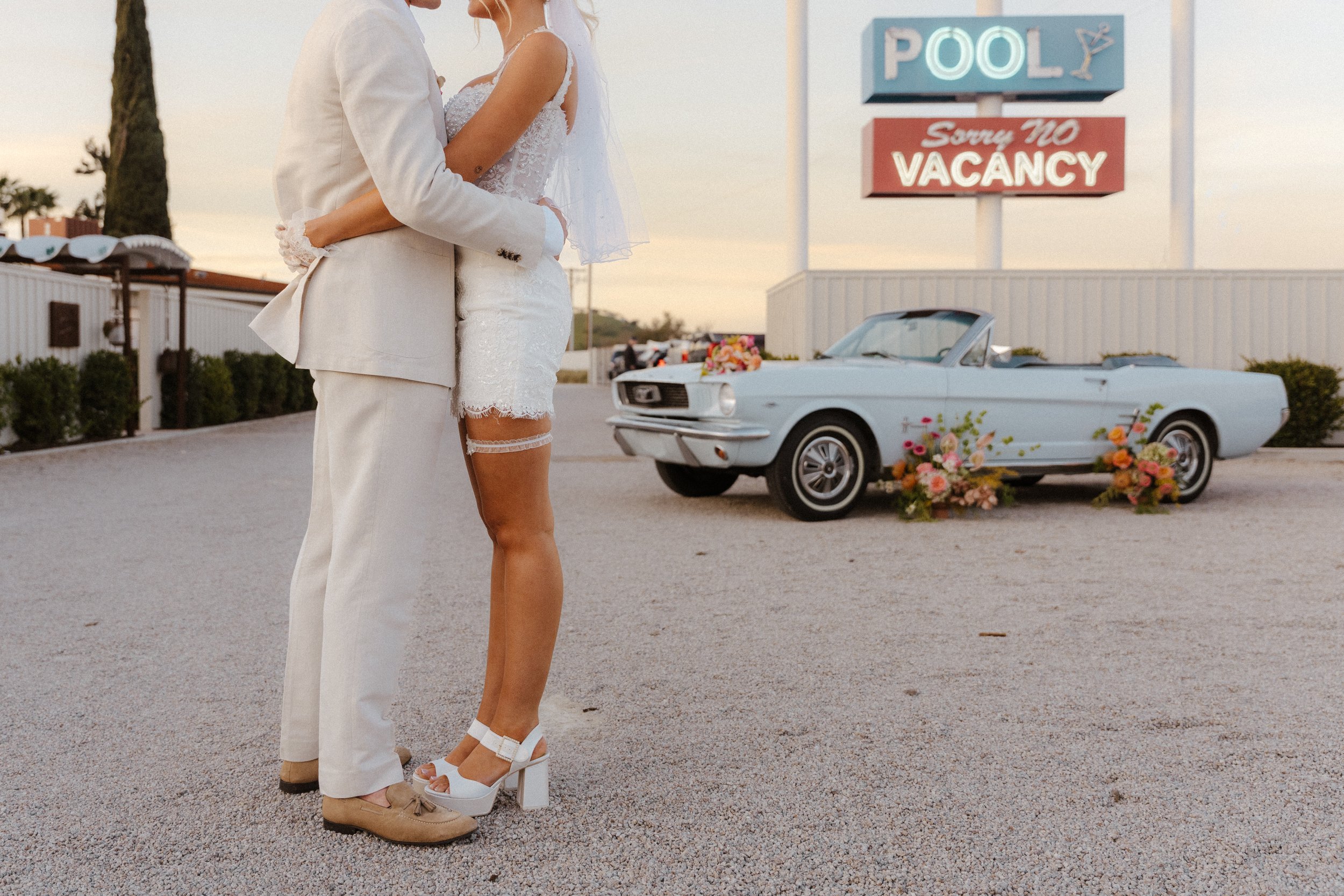 A couple stands together in a desert landscape. They're embracing in front of a baby blue classic Mustang with a hotel's neon sign in the distance.