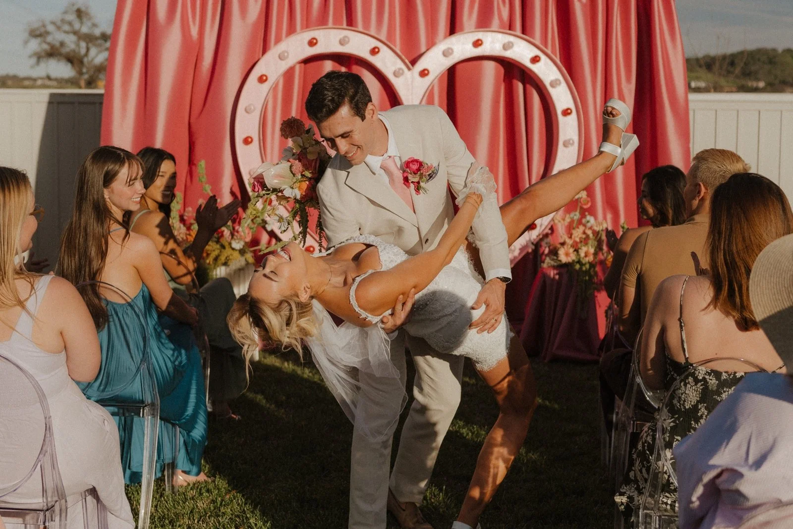 A groom dips his bride partway down the aisle. There's a pink heart arch in the background and their friends look on with smiles.