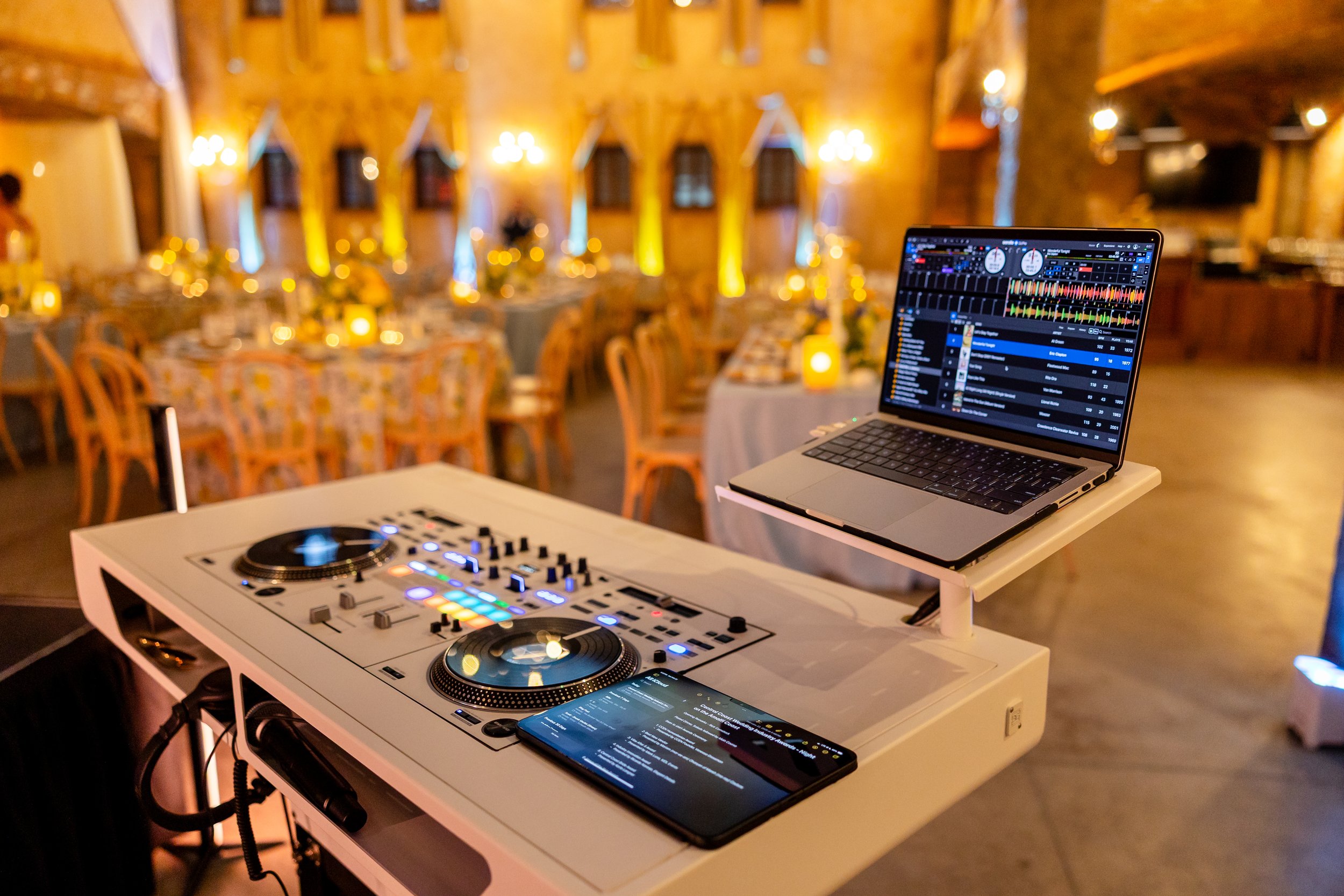 The view from behind our all white DJ podium before an awards gala begins. The setup is indoors, decorated with warm color tones, and the room is lit with yellow and blue spring colors.