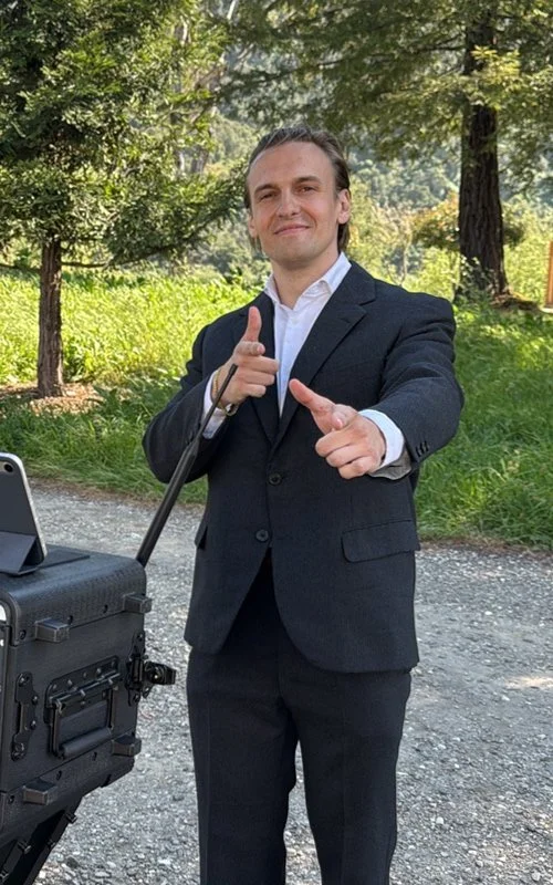 DJ Liam is outside at a wedding ceremony. He is smiling, his hair is combed back, and he's wearing a dark gray blazer with matching pants.