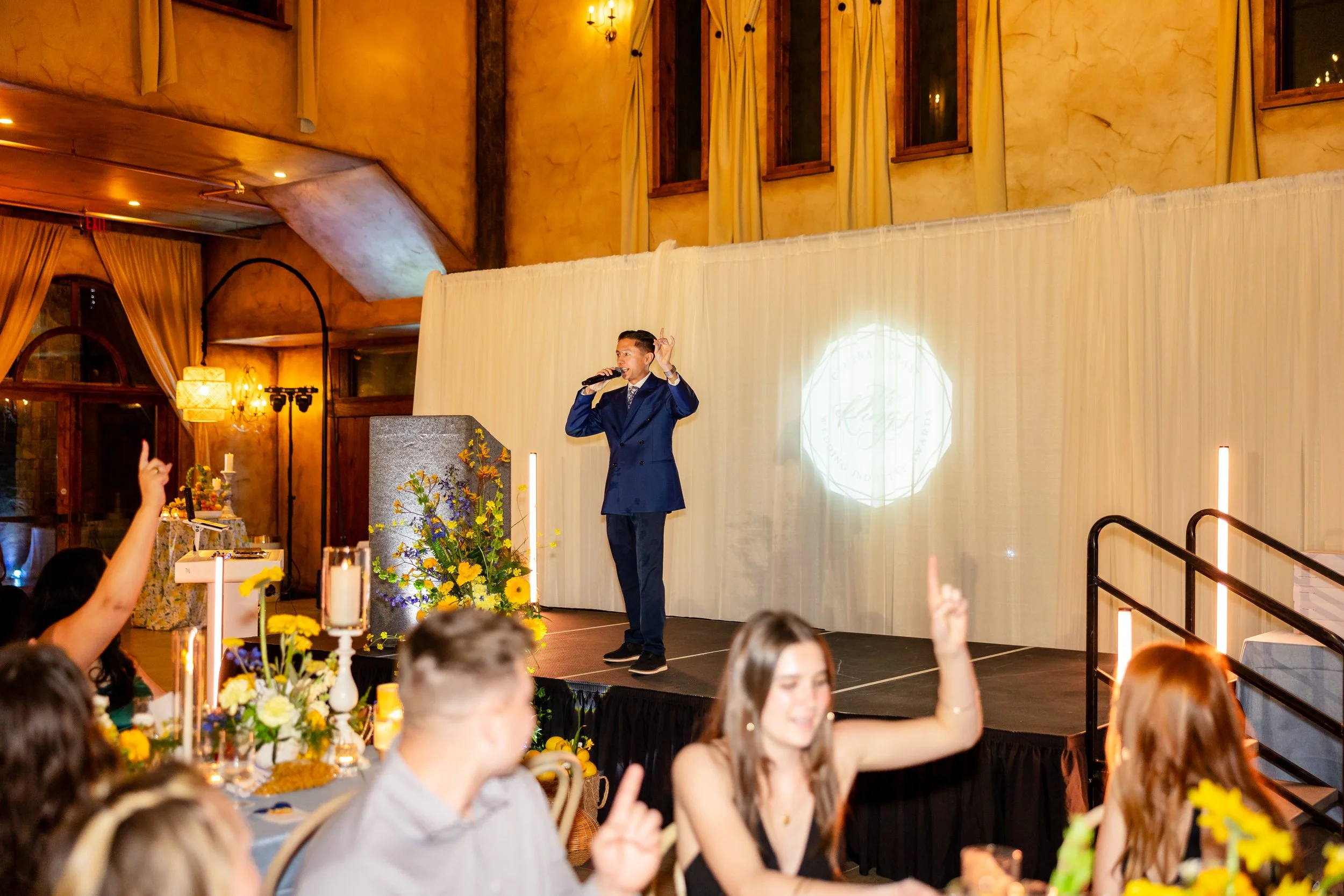 DJ Steven is standing on a stage, wearing a dark blue suit, and talking into a microphone. A logo is projected on the white draping behind him. As Steven holds up a finger, signaling "one", other event guests can be seen doing the same.
