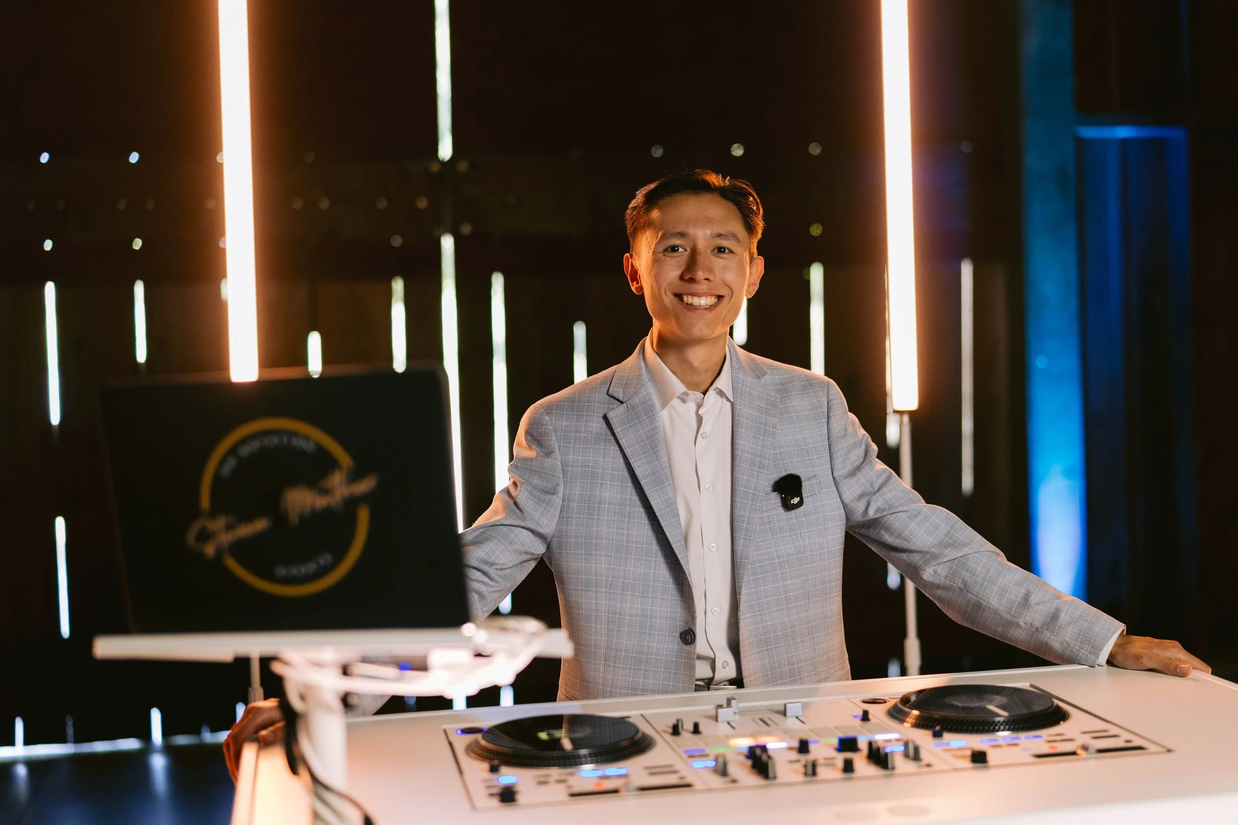 DJ Steven stands behind his all white DJ booth. He's smiling, wearing a light blue suit, and ready to DJ his next event!