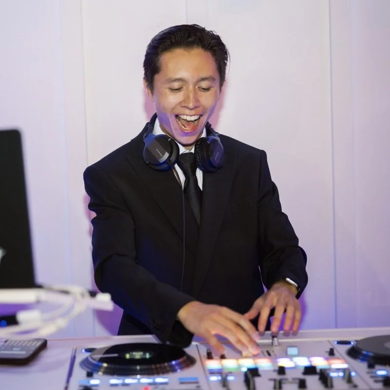 DJ Steven is behind his DJ Booth in a black suit with a black tie. He's smiling, looking down at his DJ turntables and mixing music for a dance party!