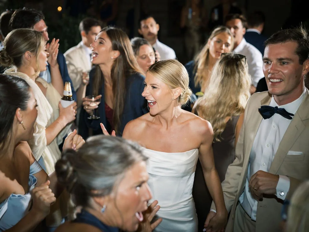 A couple is dancing at the center of their dance floor! The bride and groom are smiling, holding hands and surrounded by dancing friends and family!