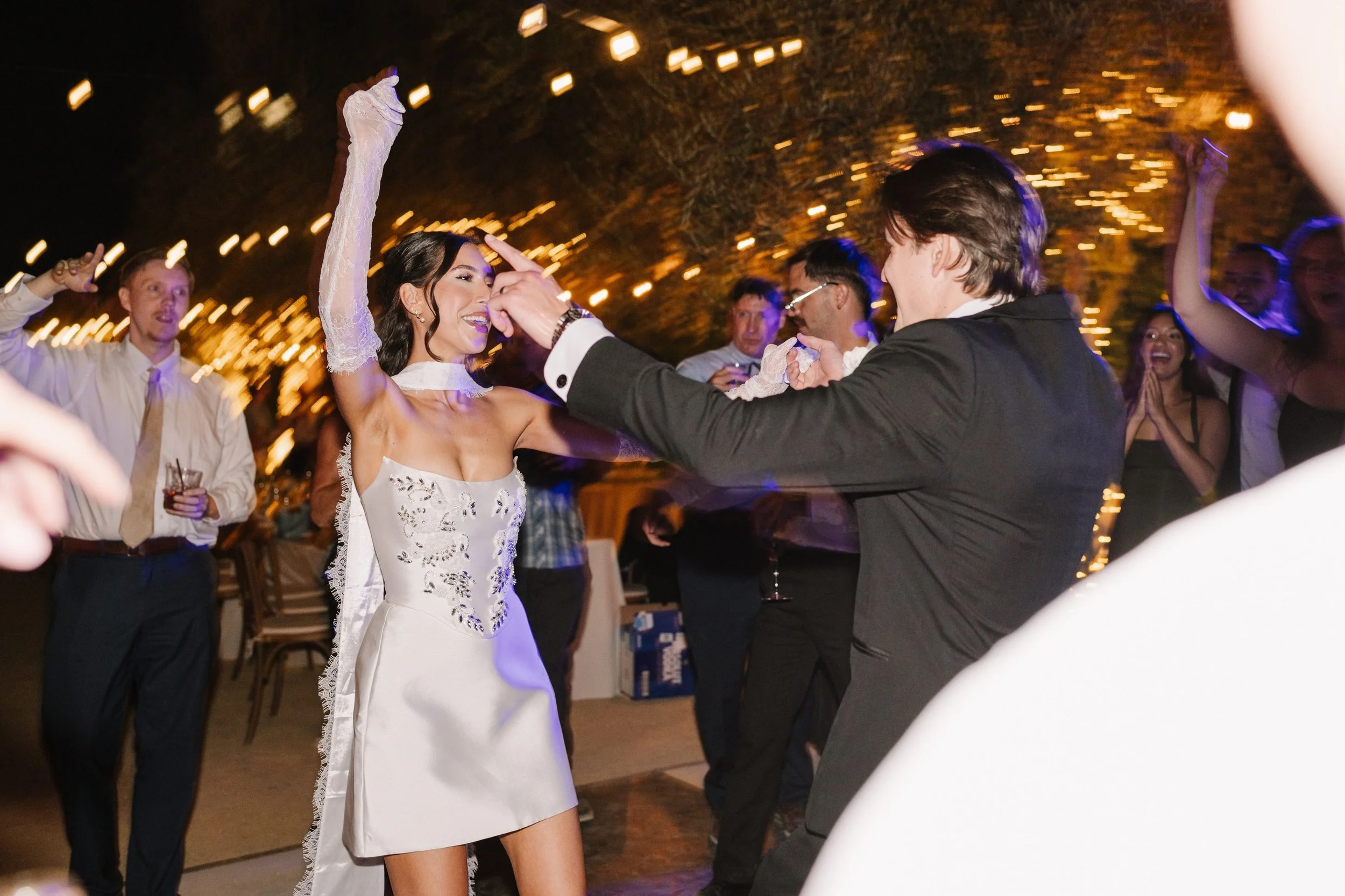 Photo Credit: Nikkels Photography, Allegretto Resort, Paso Robles Wedding (A bride and groom dance together on their wedding night. Their friends and family surround them. Many guests have their hands in the air, are smiling, and happy for the newlyw