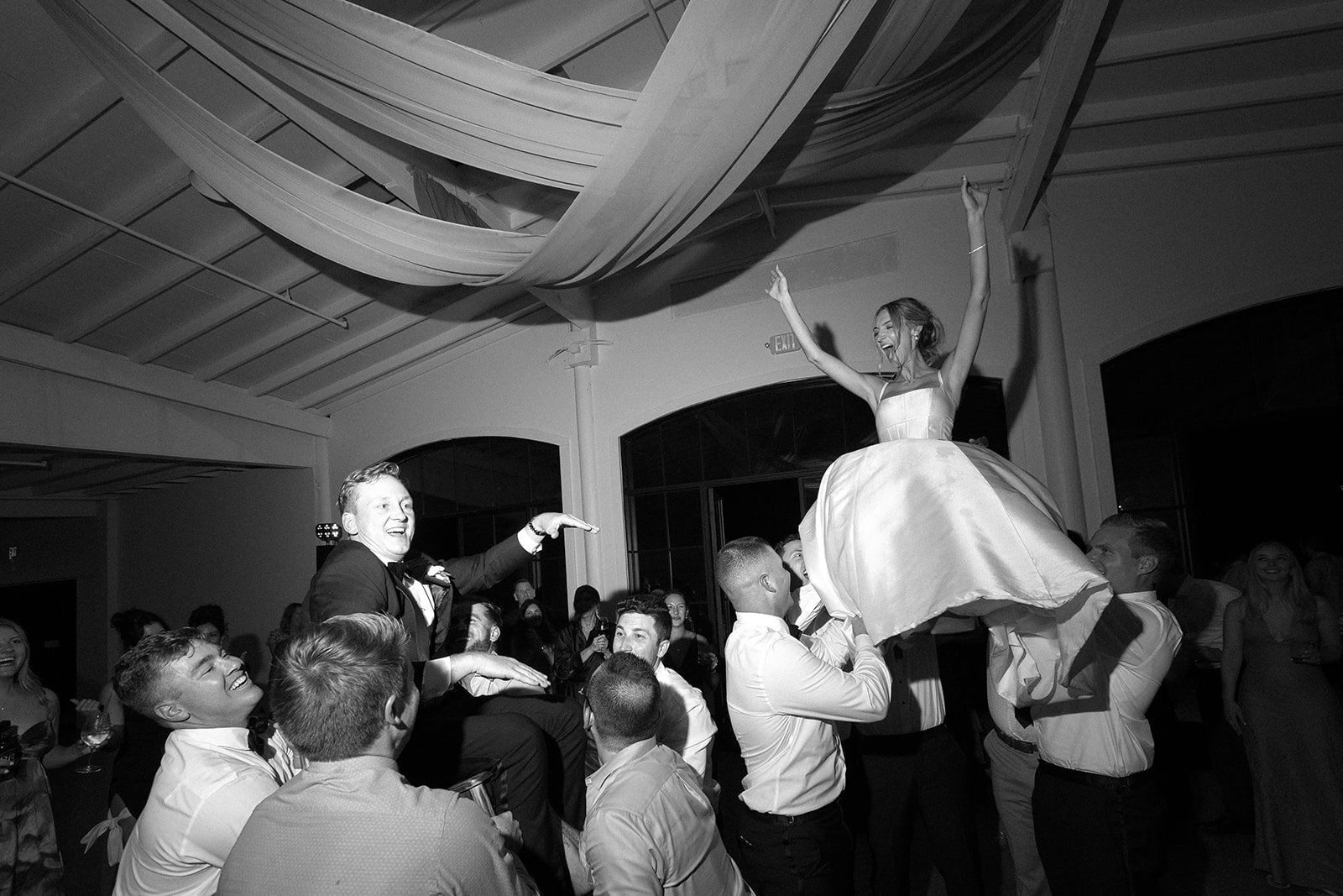 A bride and groom are hoisted into the air by their friends and family on the dance floor! This moment will be unforgettable! The groom is smiling with a hand in the air. The bride is screaming with joy and has both hands in the air!