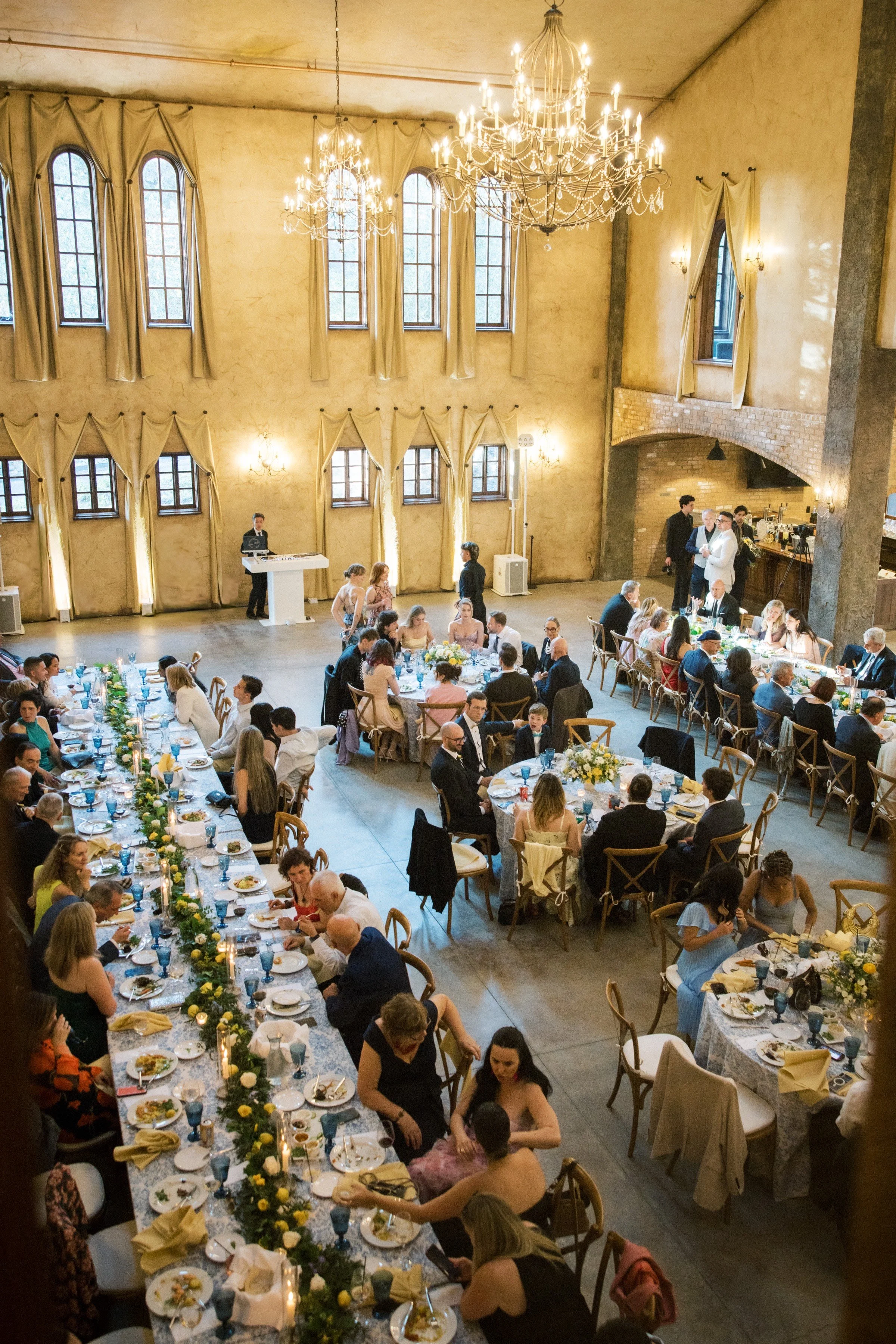 DJ Steven stands behind his white DJ podium inside the wedding reception hall at Villa Loriana, a private estate, with Italian style architecture. Wedding guests are seated and talking at dinner tables adorned with blue and green accents.