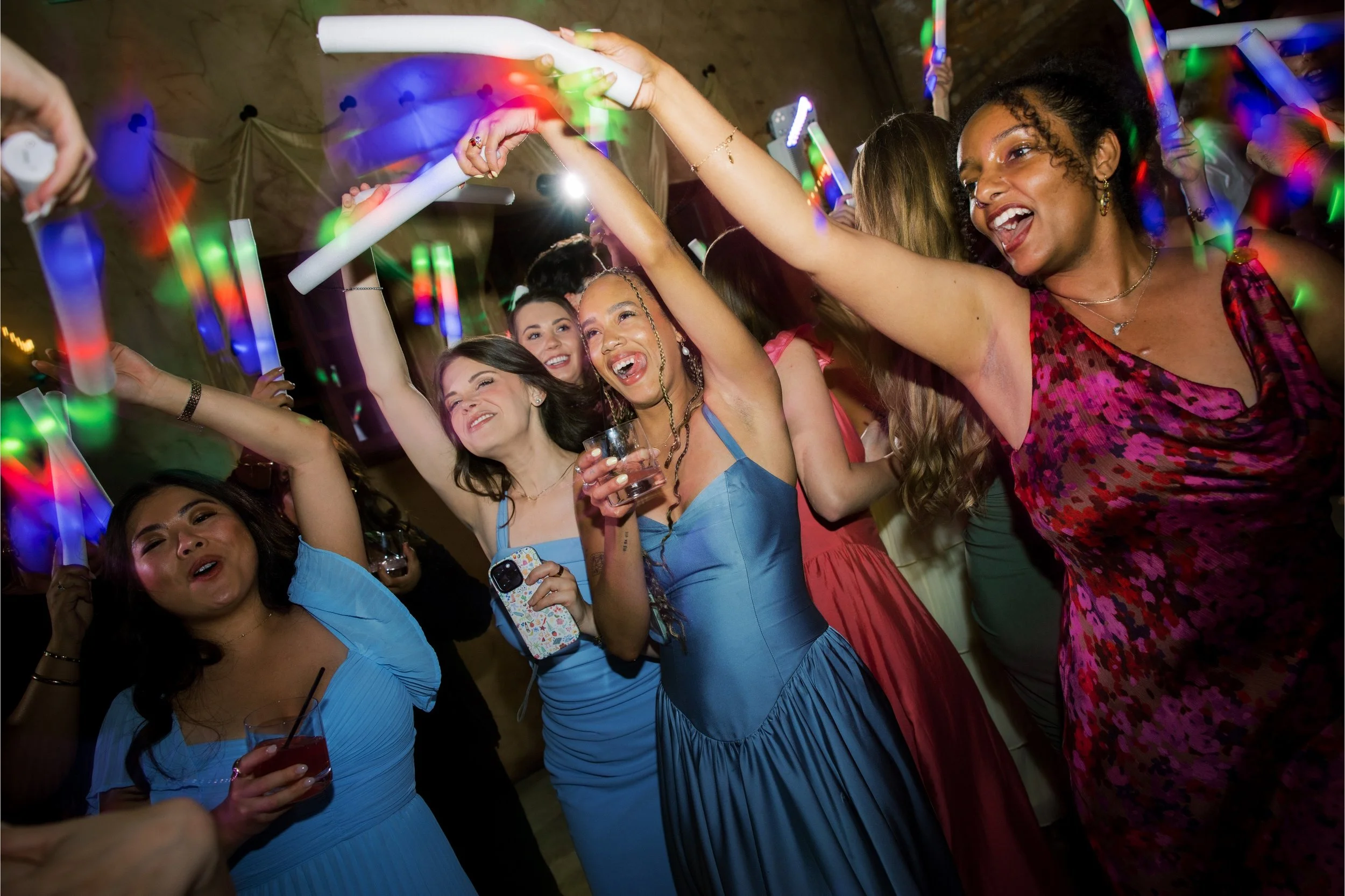 Photo Credit: Ashley Rae Collective, Villa Loriana, San Luis Obispo Wedding (A couple's bridesmaids and friends are dancing together. Everyone is holding an LED foam stick and the bridesmaids are wearing blue dresses. Many people are smiling in this 