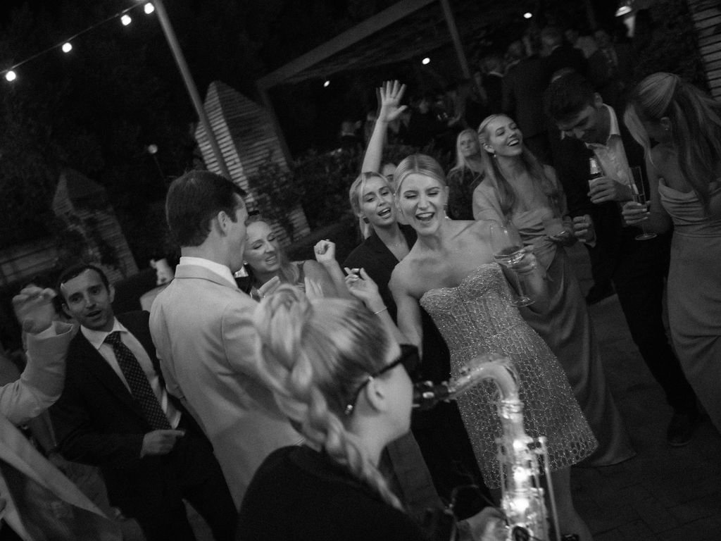 A bride and groom dance together surrounded by friends and family on their wedding night. A female saxophonist can be seen playing in the foreground.