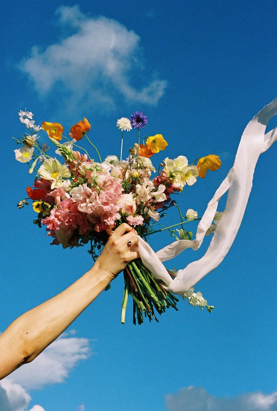 Bride's hand raising a colourful wildflower bouquet against a blue sky, photographed by Georgia Lilley