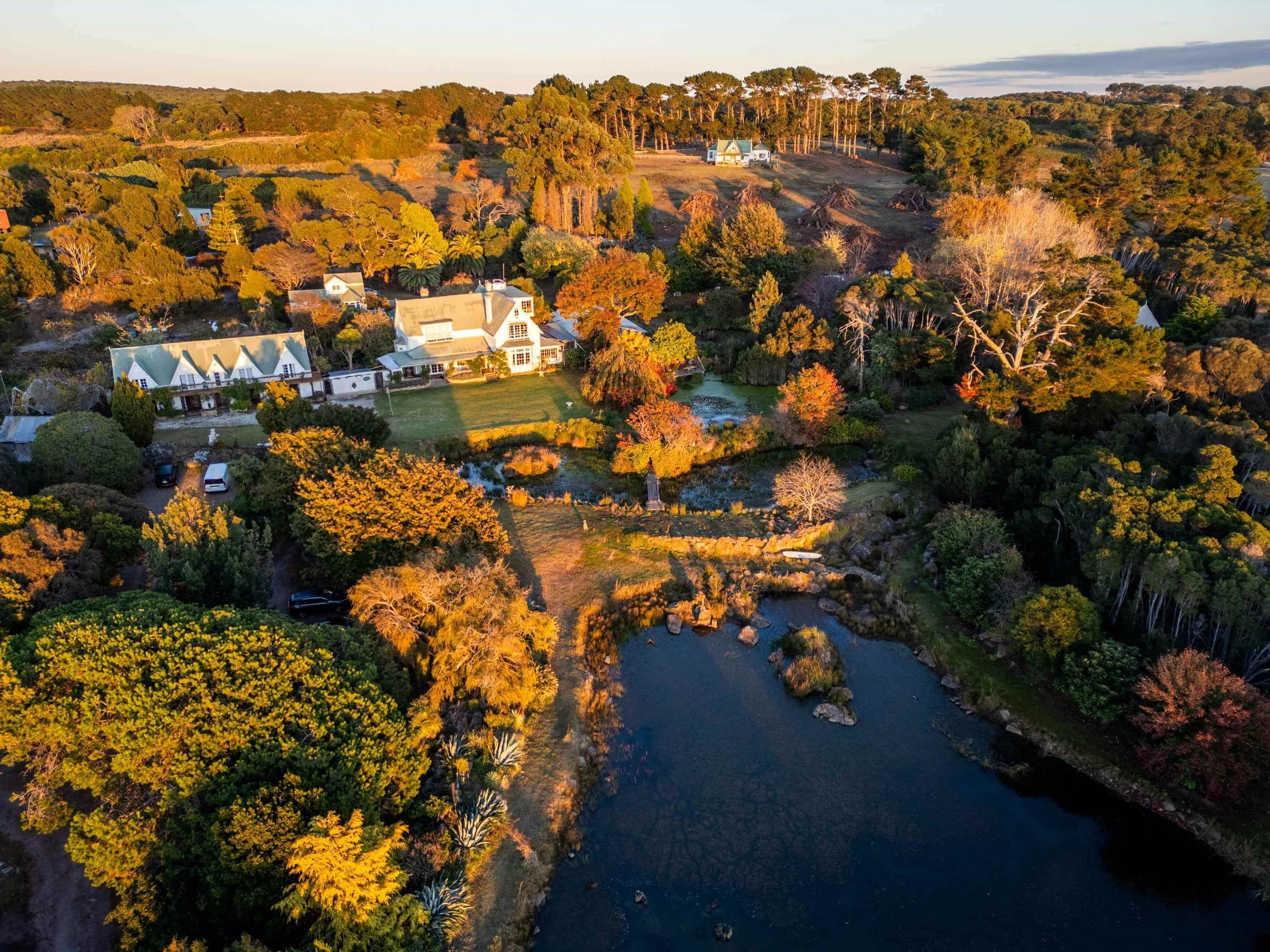 Golden hour aerial view of Hawley House wedding venue, showing the heritage homestead, gardens and dam surrounded by autumn foliage on Tasmania's north west coast