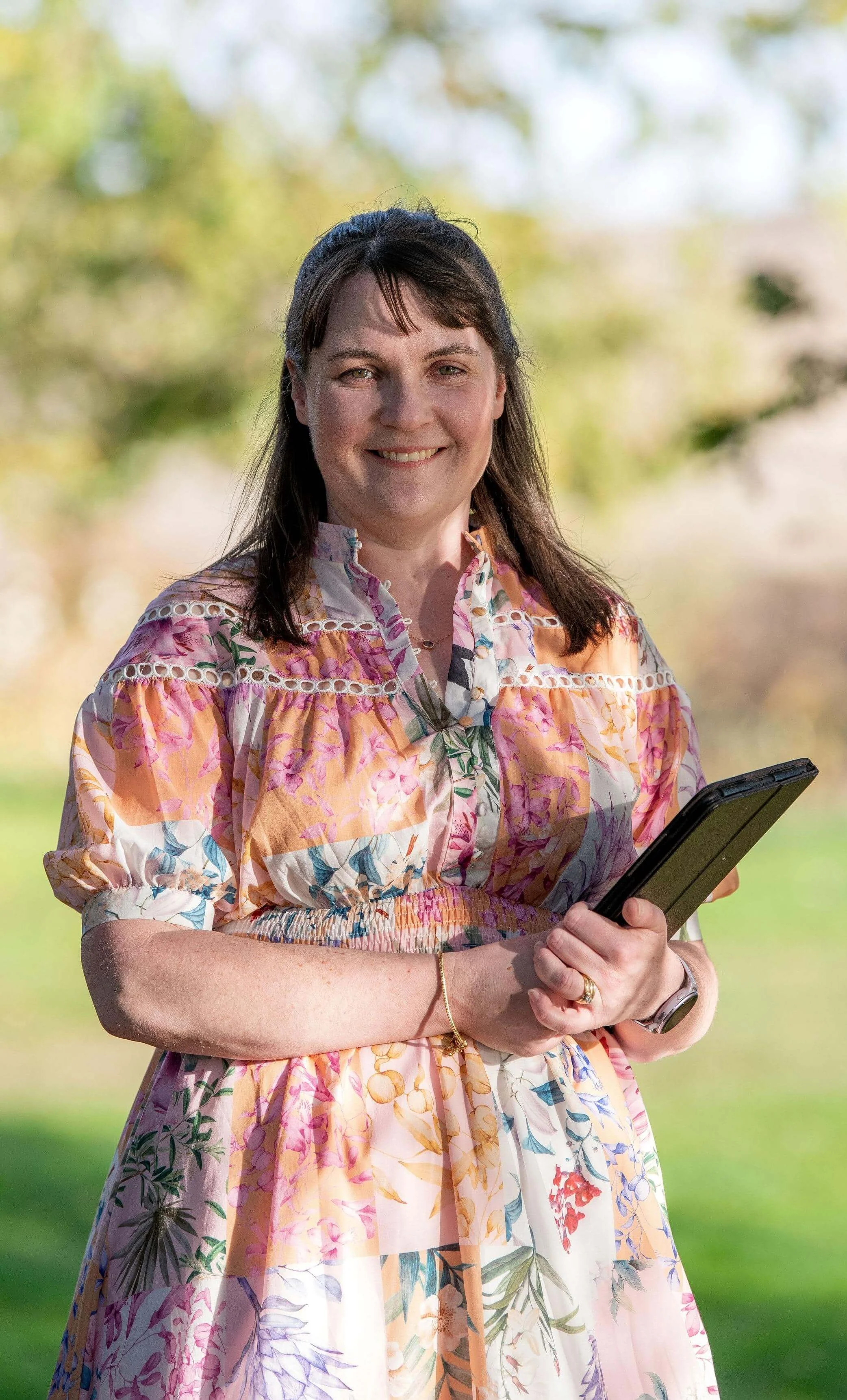 Claire Archer, Tasmanian wedding celebrant, smiling outdoors in a floral dress holding a ceremony folder