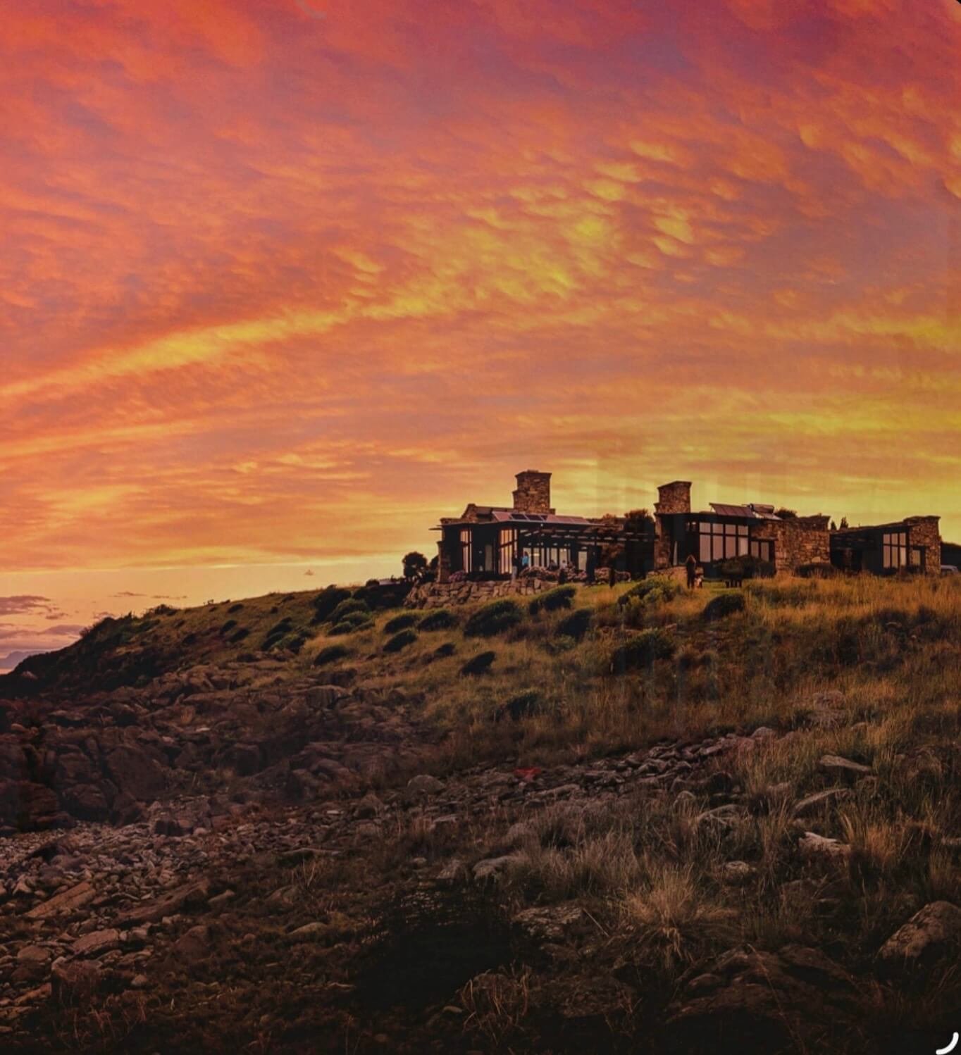 Stone and timber lodge on a coastal Tasmanian headland at sunset, silhouetted against a dramatic orange sky — Thalia Haven elopement accommodation