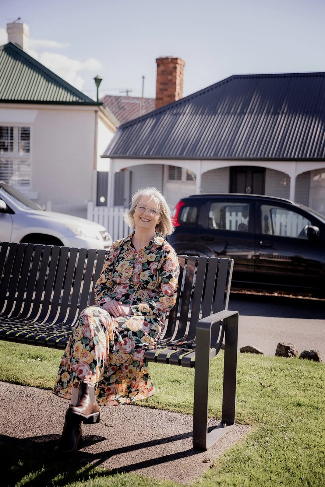 Mary Figg, Hobart marriage celebrant, smiling on a park bench in Battery Point wearing a floral dress