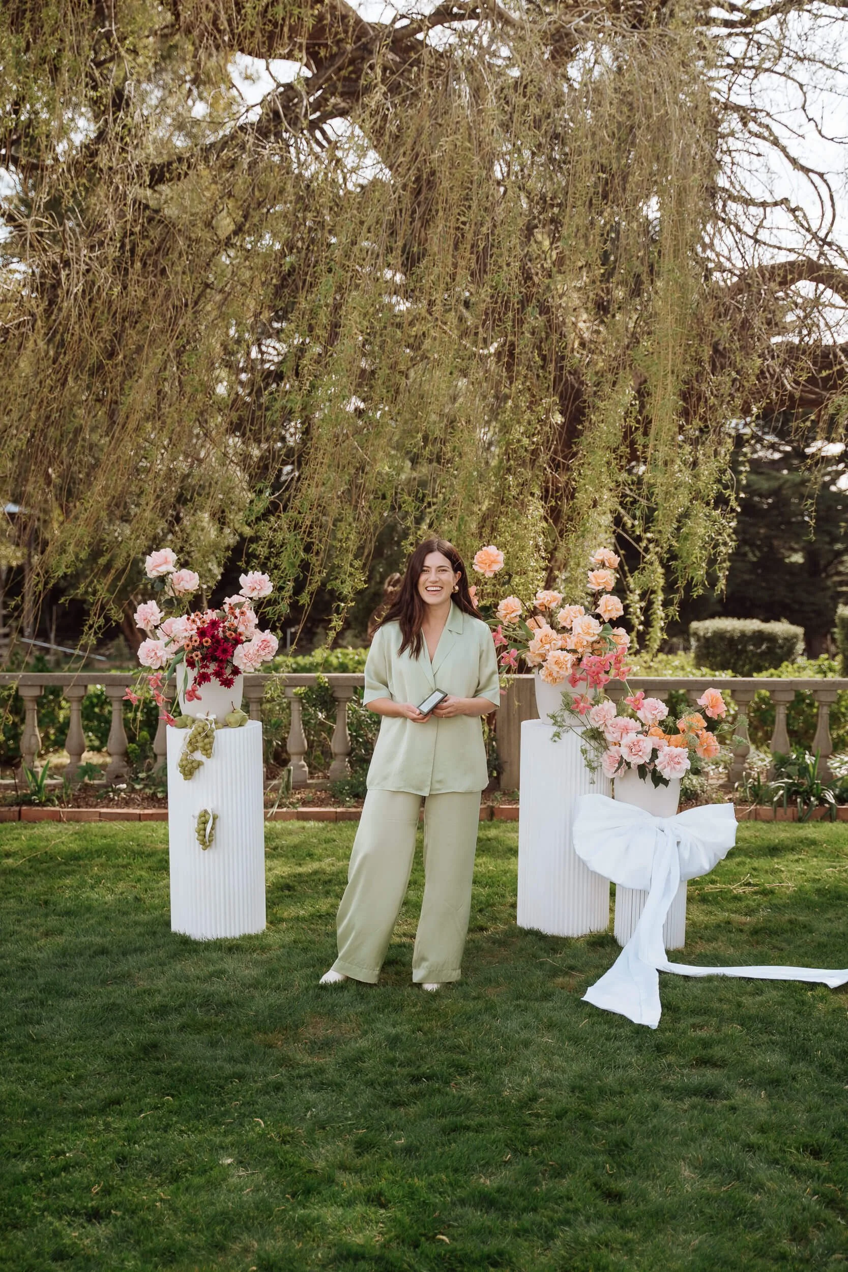 Celebrant standing between bountiful flowers and plinths, smiling and wearing green silk suit 