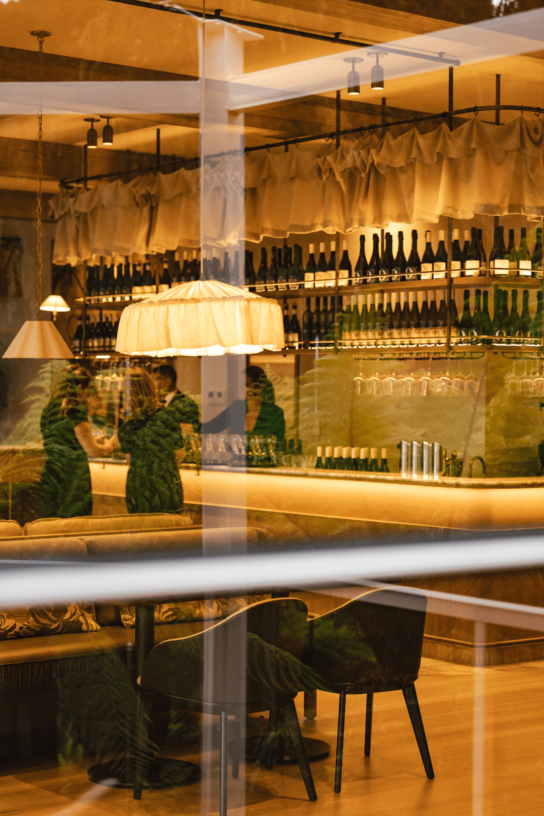 Warm-lit wine bar interior at Bird in Hand, Royal Tasmanian Botanical Gardens, viewed through glass with bottle-lined shelves and lounge seating