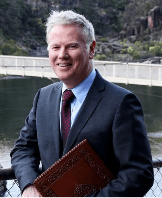 John Burke, Tasmanian marriage celebrant, smiling in a suit beside a river gorge holding a ceremony folder